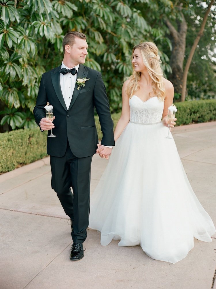 A newlywed couple walking hand in hand outdoors, smiling and holding glasses with drinks, on a pathway with green foliage in the background.
