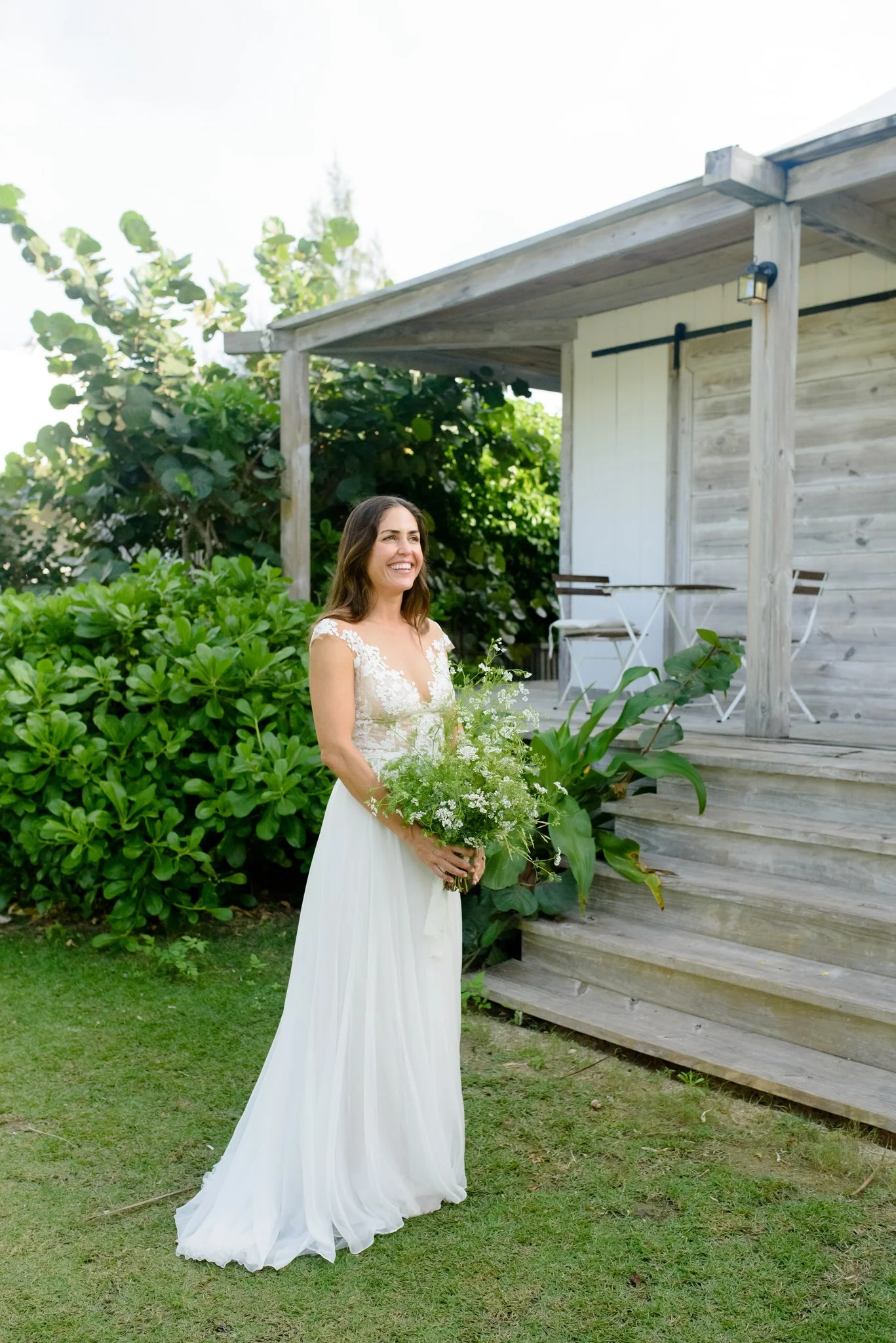 A woman in a white wedding dress holding a bouquet of white and green flowers, standing outdoors near a wooden deck and lush green bushes at The Other Side Hotel Eleuthera during a luxury Bahamas destination wedding.