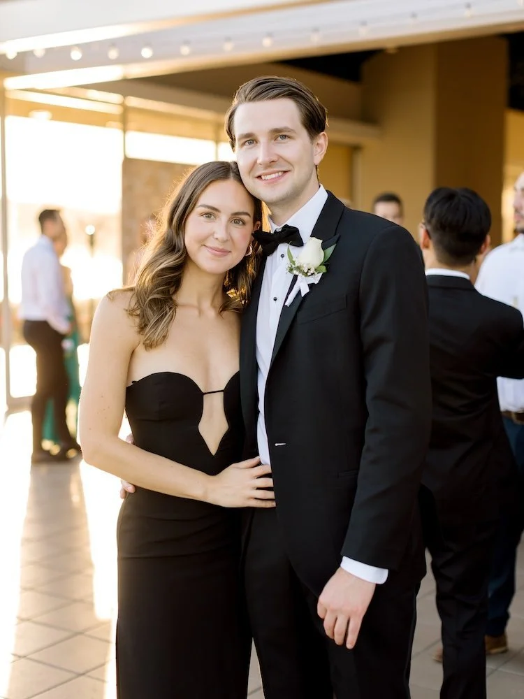 A young couple dressed in formal attire, the woman in a strapless black dress and the man in a tuxedo, smiling and posing at a social event or wedding reception, outdoors with other guests in the background.
