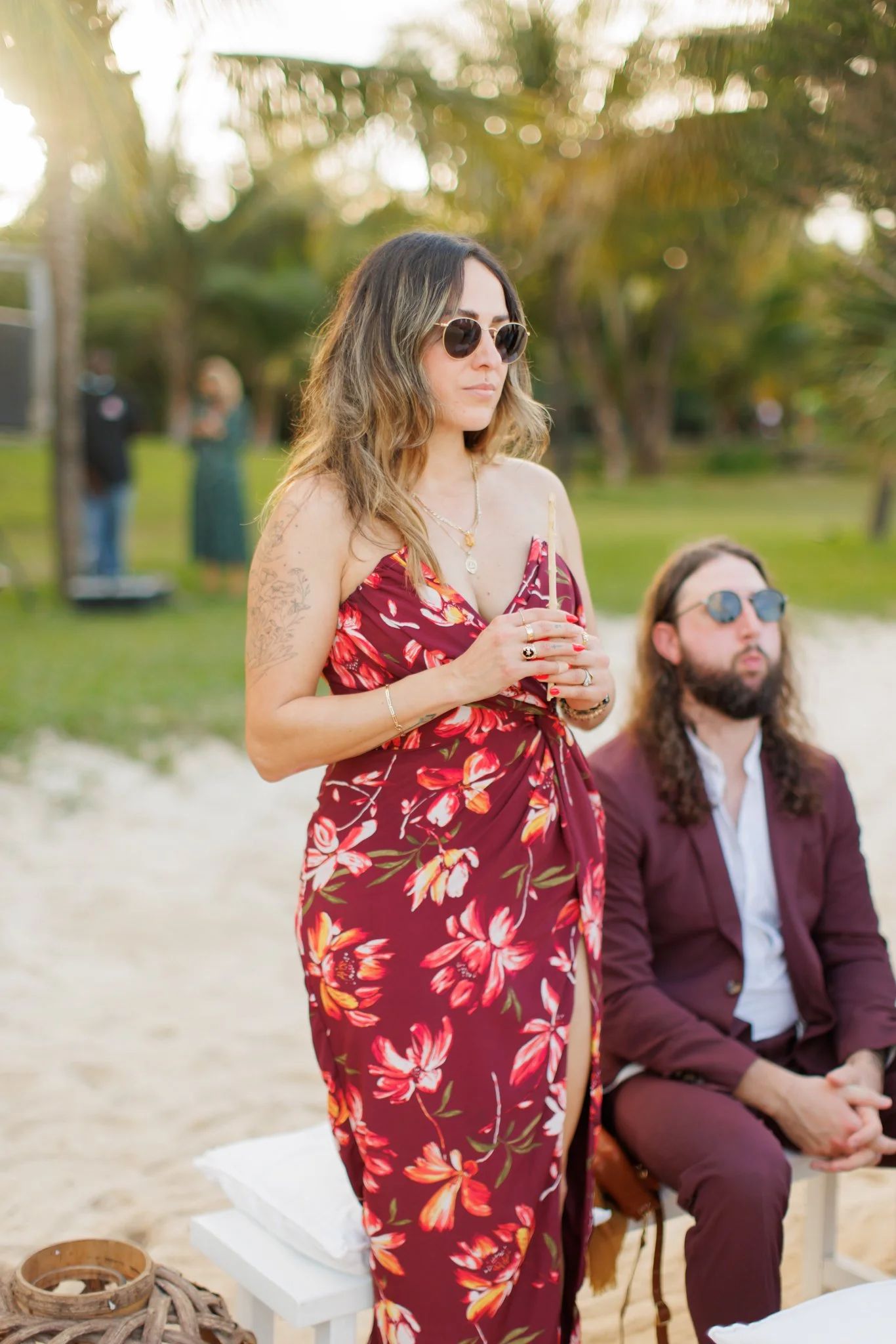A woman in a red floral dress with sunglasses and jewelry holds a candle during an outdoor ceremony, with a seated man in a burgundy suit nearby, at a beach setting with palm trees.