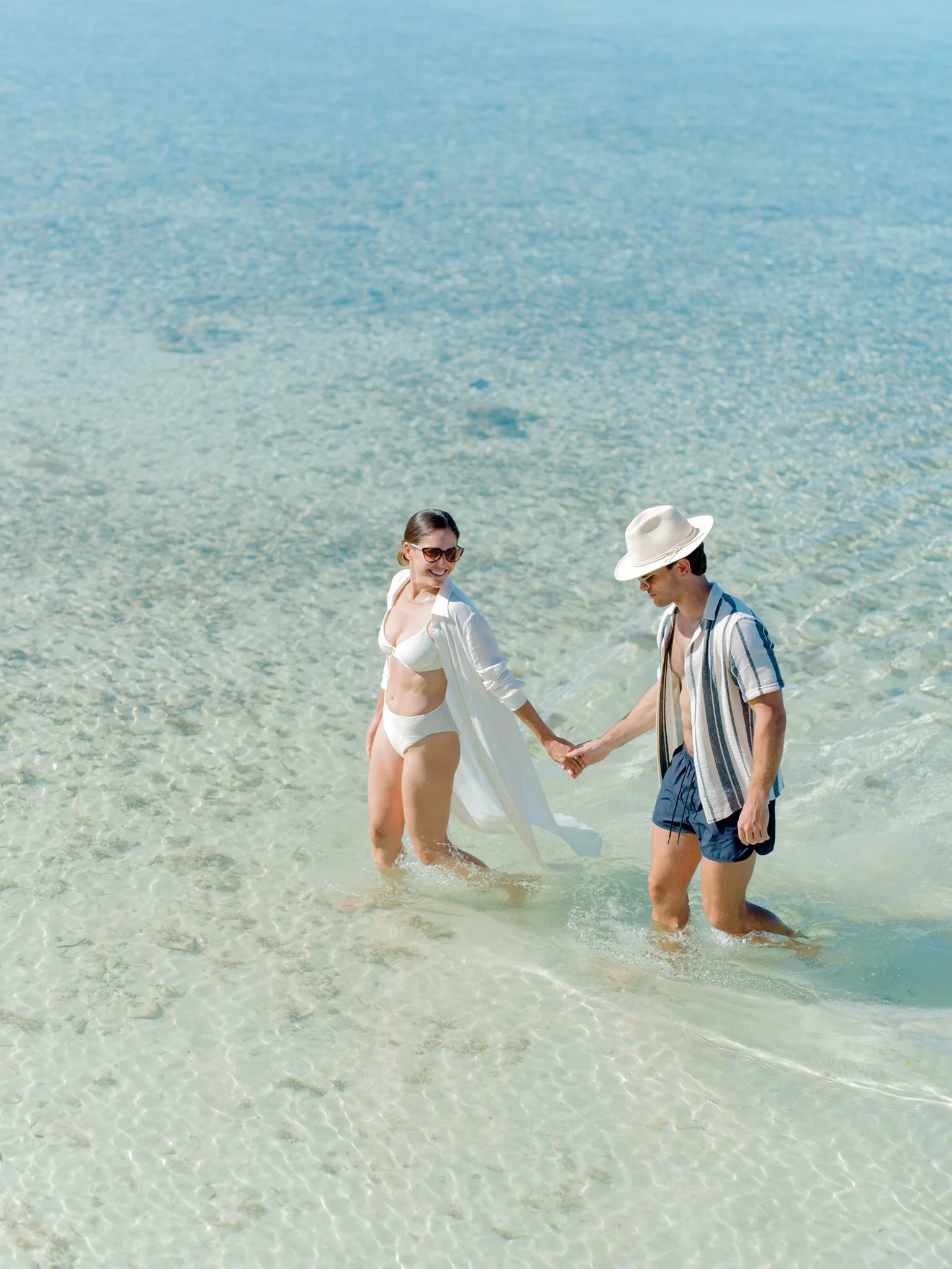 A couple holding hands and walking through clear, shallow water at the beach at The Other Side Hotel during their destination wedding in Eleuthera, Bahamas.