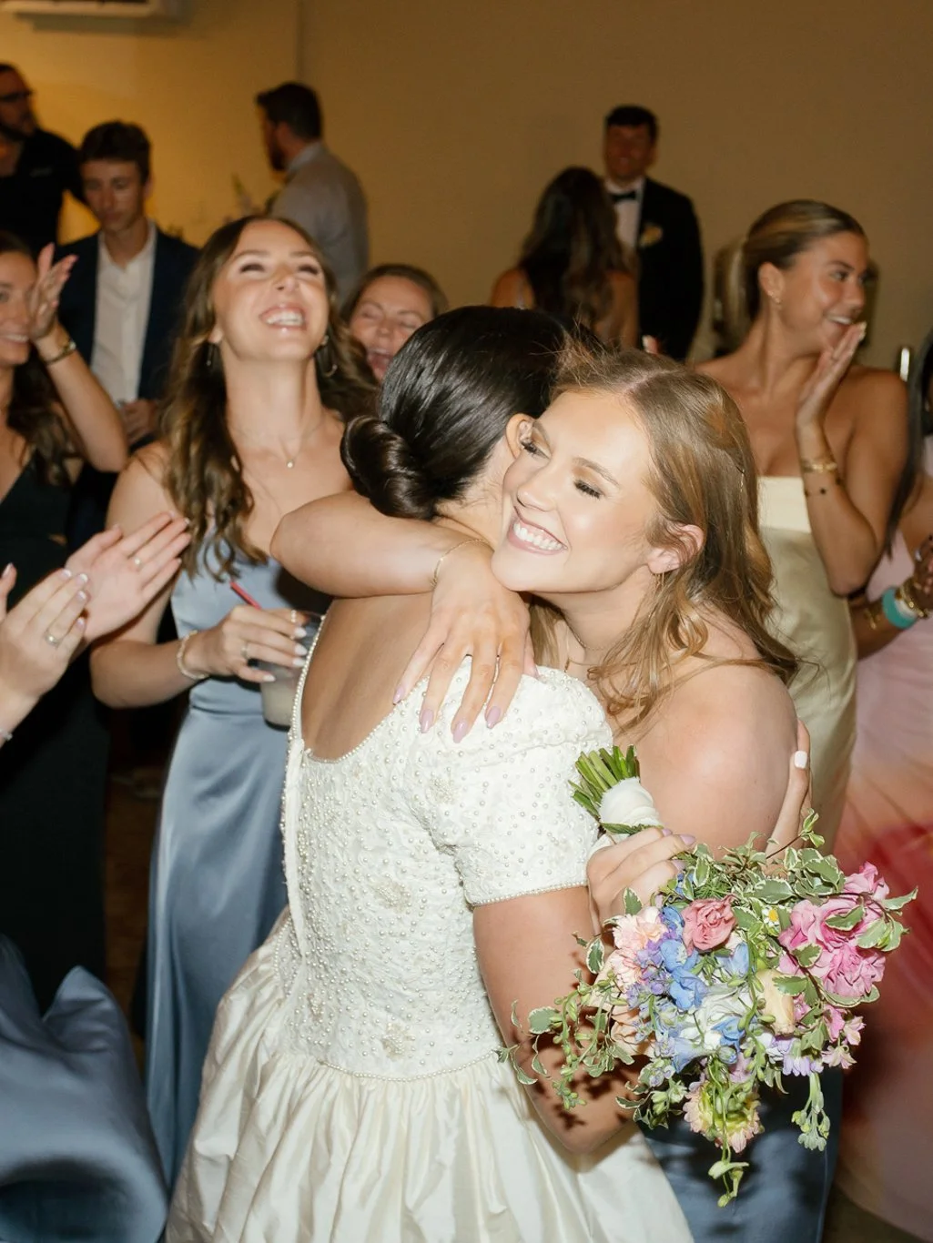 Two women hugging at a wedding reception, one in a white wedding dress holding a bouquet of flowers, surrounded by smiling guests.