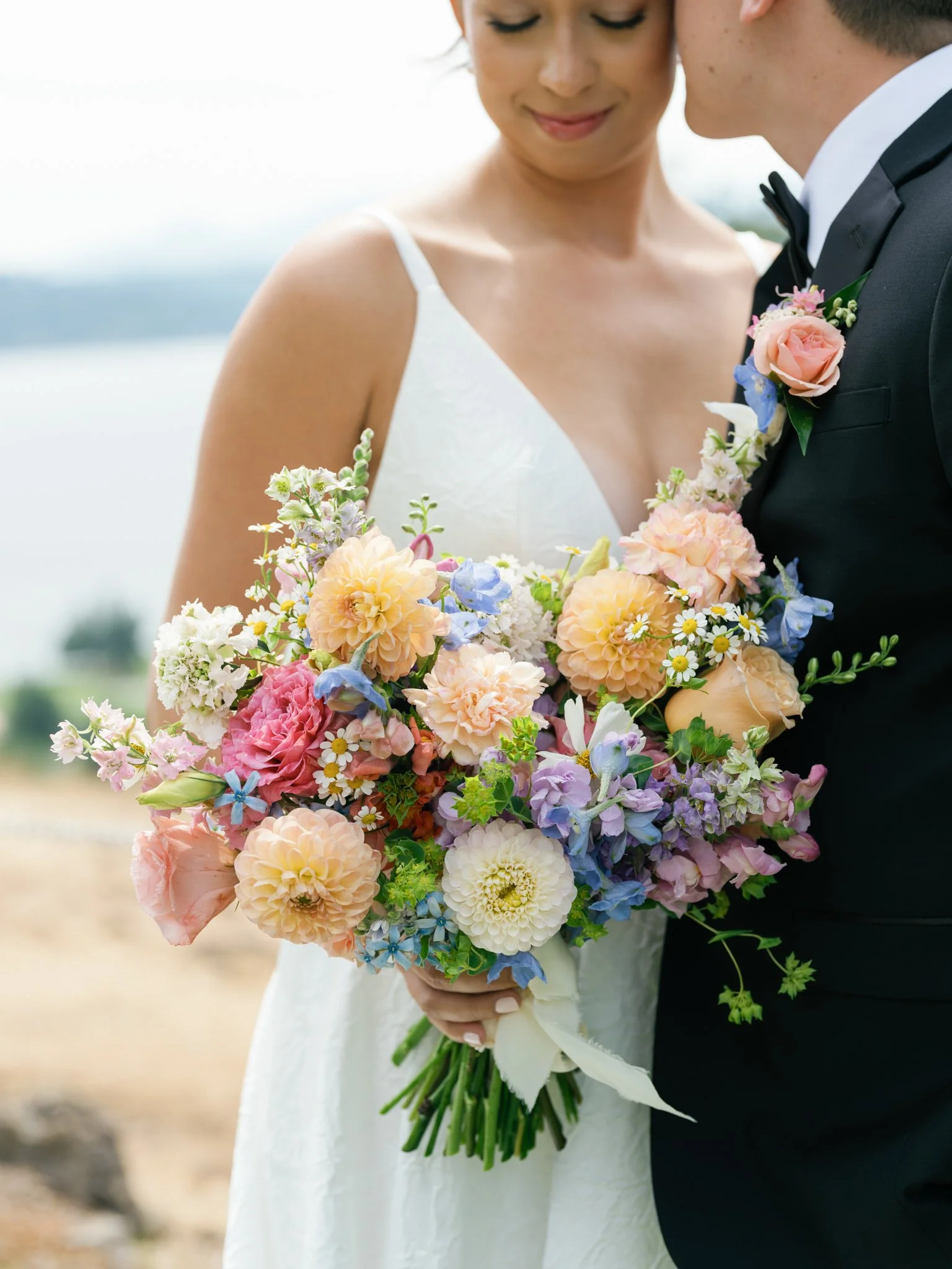 Bride holding a colorful bouquet of flowers while standing close to groom in a black tuxedo, outdoors by a body of water in North Idaho.
