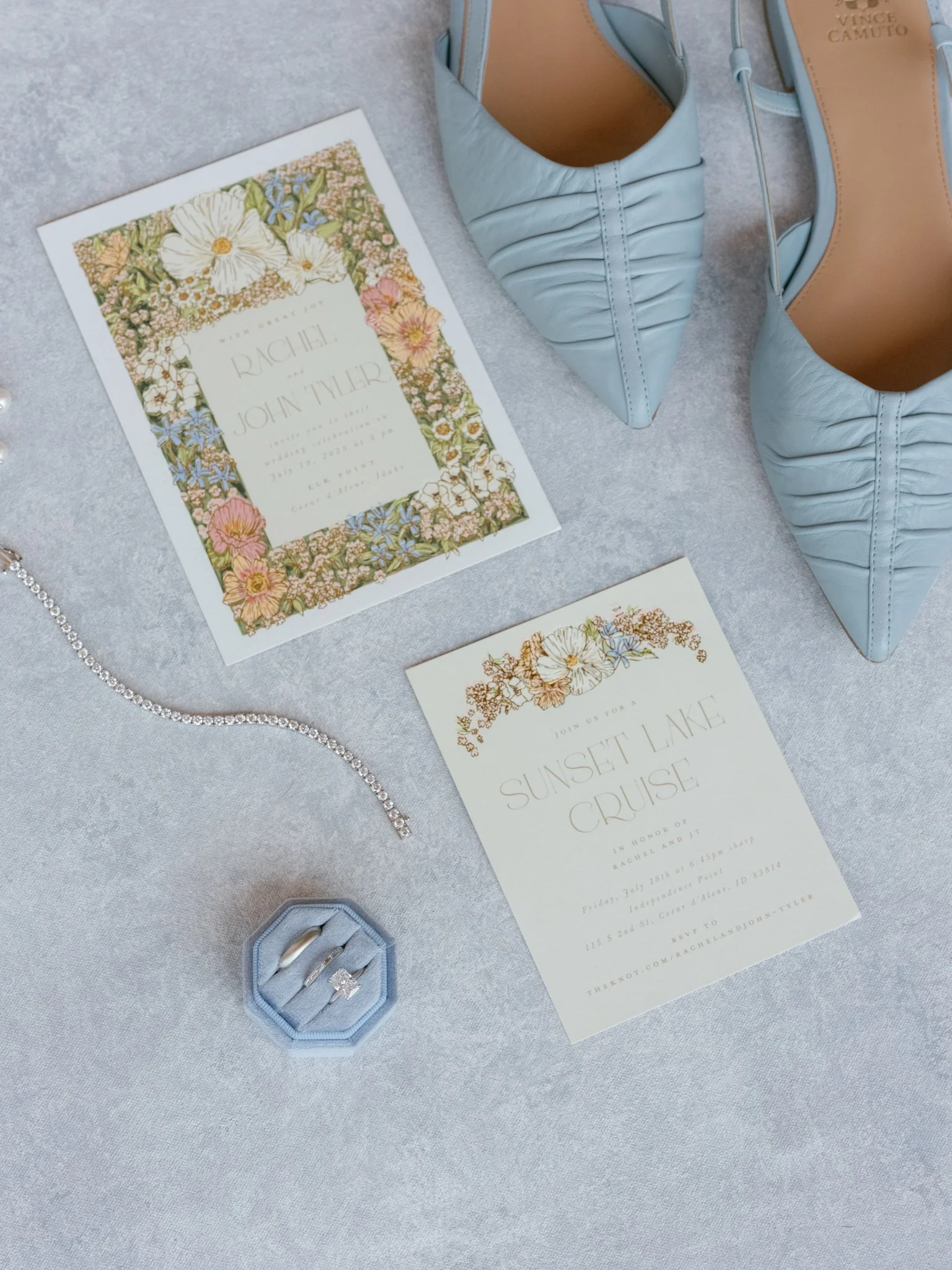 Light blue pointed-toe heels, a wedding invitation with floral border for Rachel and John Tyler, a smaller invitation for a Sunset Lake Cruise, a diamond ring in a blue ring box, and a silver bracelet on a light gray surface.