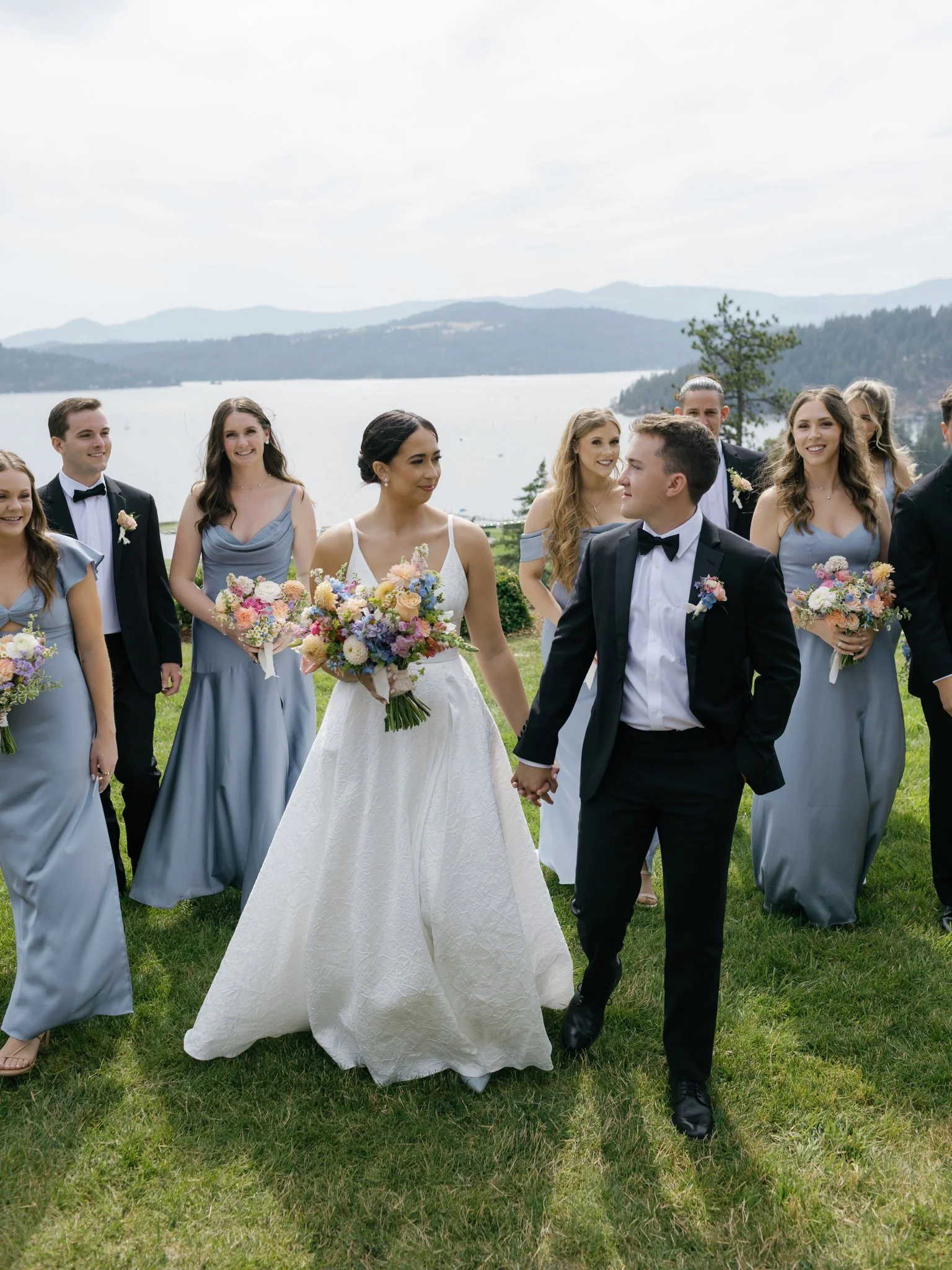 A bride and groom holding hands, walking on grass with friends during a wedding outdoors, with a lake and mountains in the background at Elk Point wedding in Coeur d’Alene.