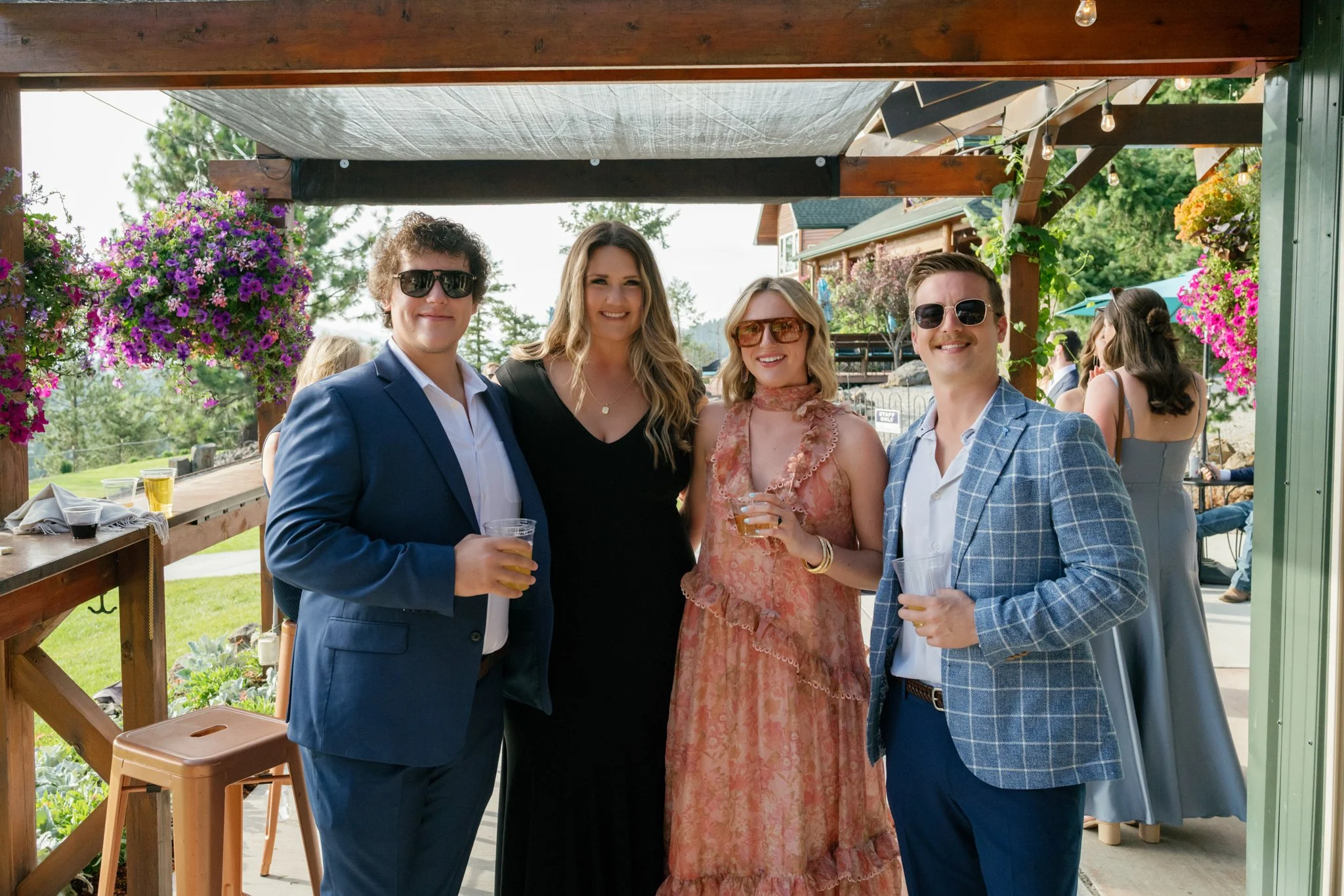 Four people dressed in formal attire with drinks in hand, standing under a wooden canopy decorated with hanging flowers, at an outdoor event.