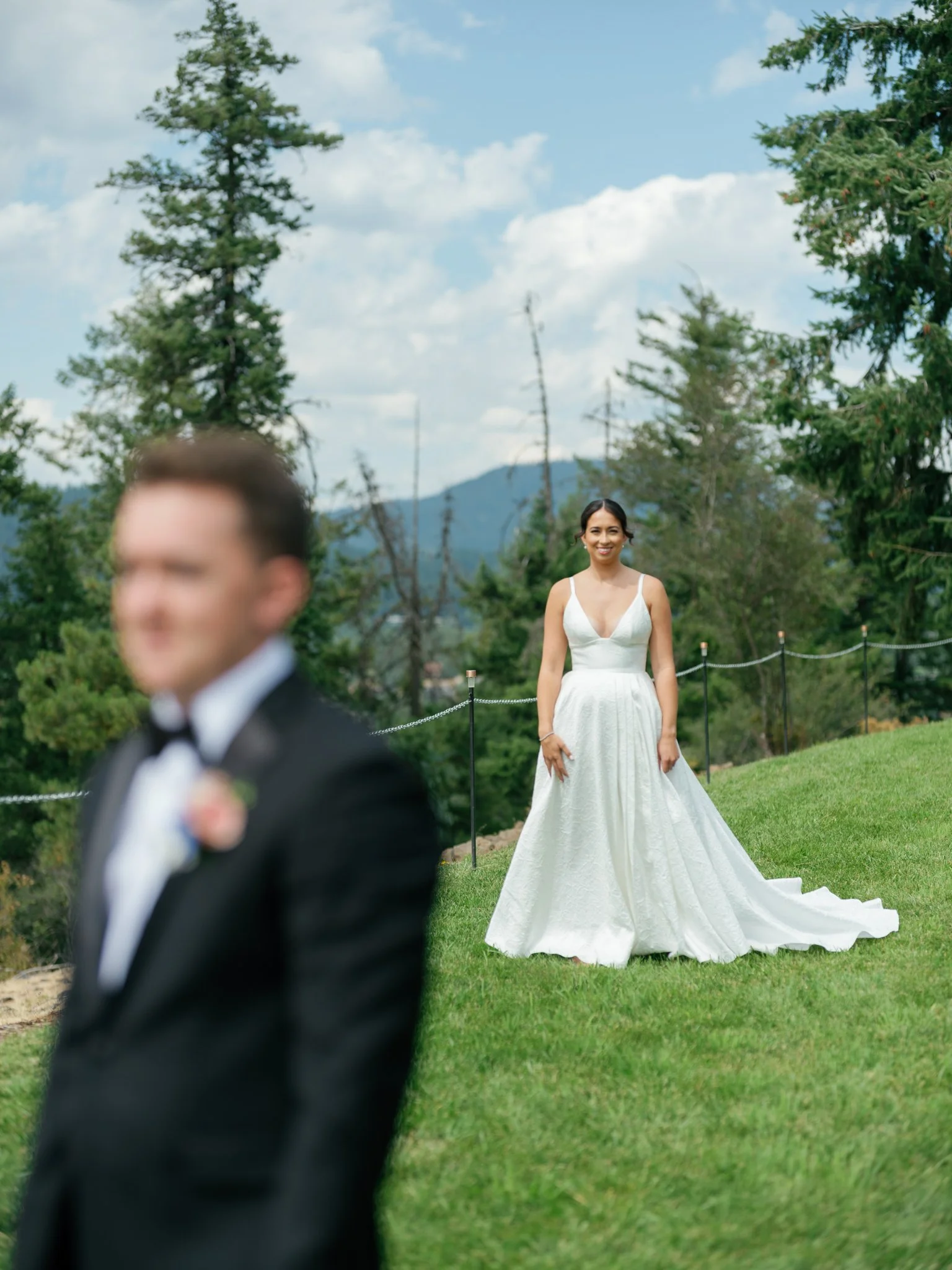 Bride in a white wedding dress smiling in the background with groom in a black tuxedo blurred in the foreground during an outdoor wedding at Elk Point wedding above Lake Coeur d’Alene.