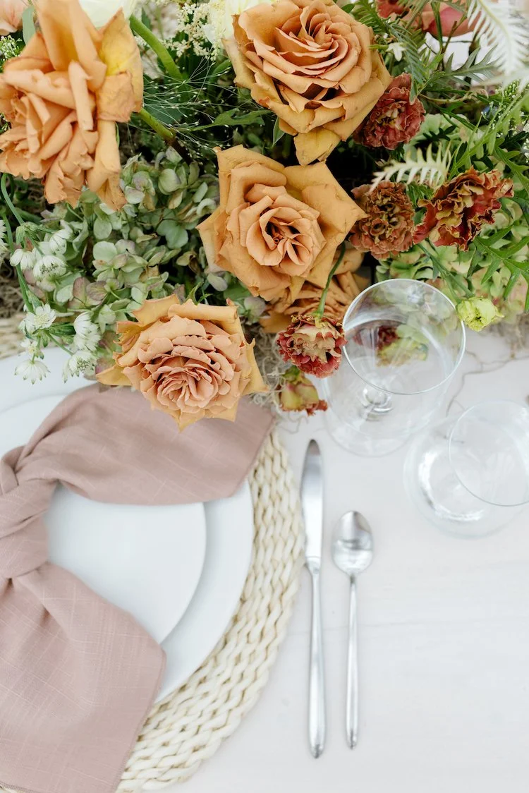 A floral table arrangement with beige roses, green foliage, and small white flowers. The table setting includes a white plate with a beige cloth napkin, silverware, and empty wine glasses.