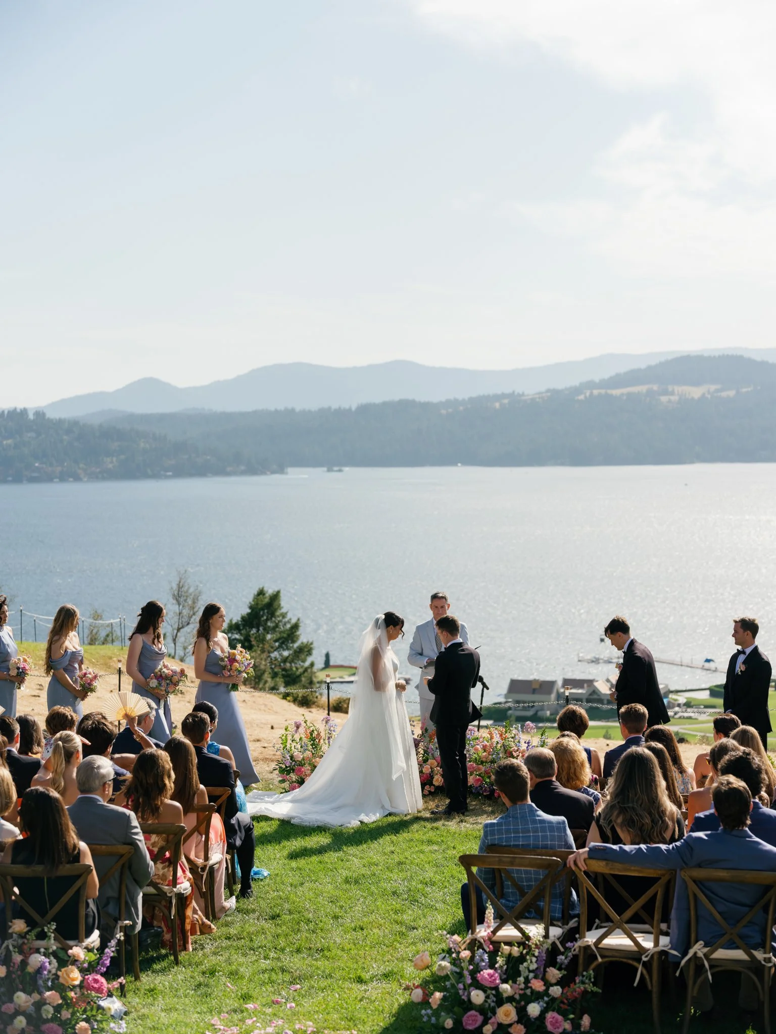 Outdoor wedding ceremony by a lake with mountains in the background, bride and groom exchanging vows, bridesmaids in purple dresses, groomsmen in dark suits, seated guests, floral arrangements, sunny weather at Elk Point wedding above Lake Coeur d’Al