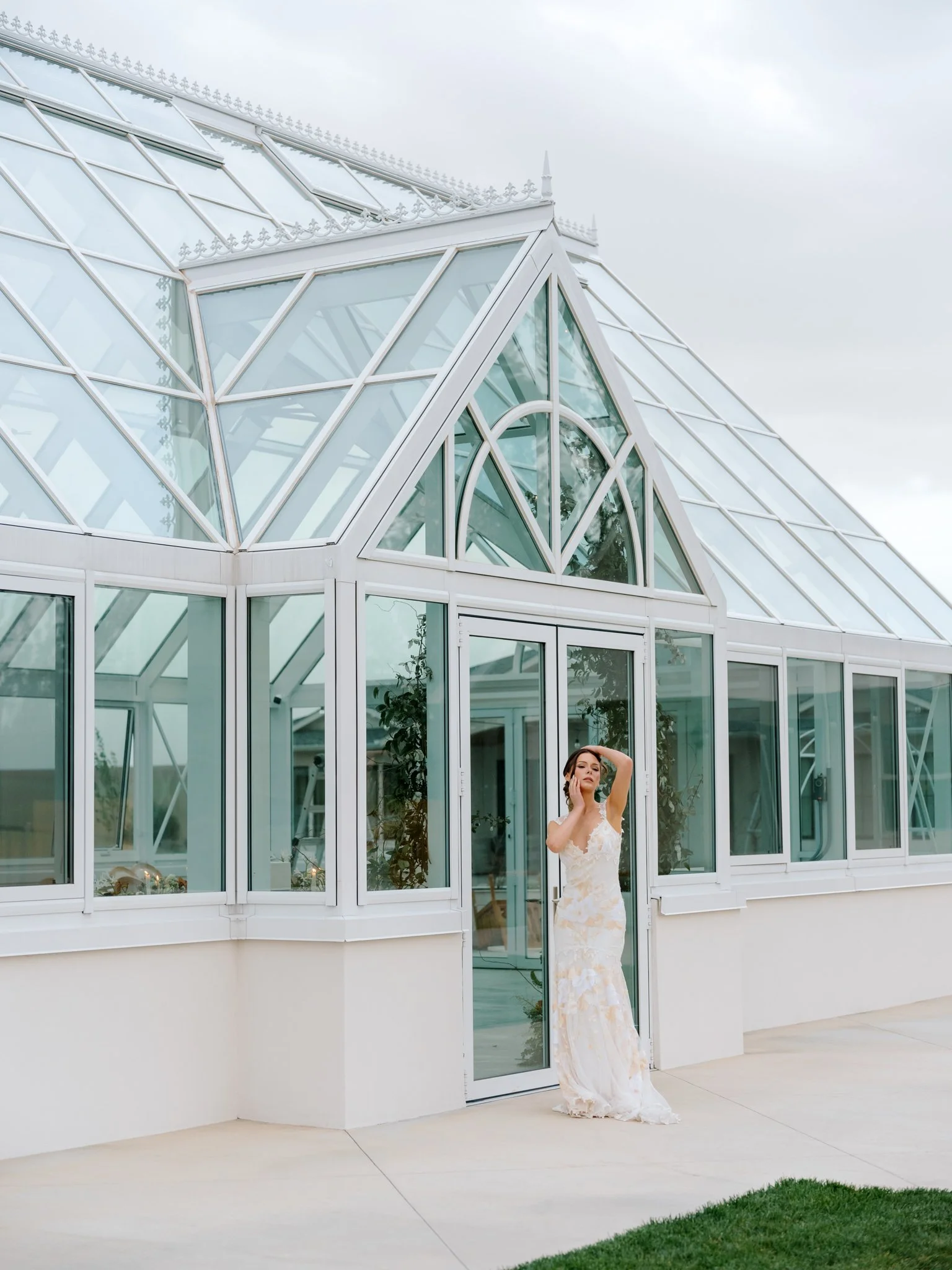 A woman in a wedding dress stands outside a modern glass greenhouse, touching her hair with one hand at Greenhouse on Harvard near Spokane, Washington