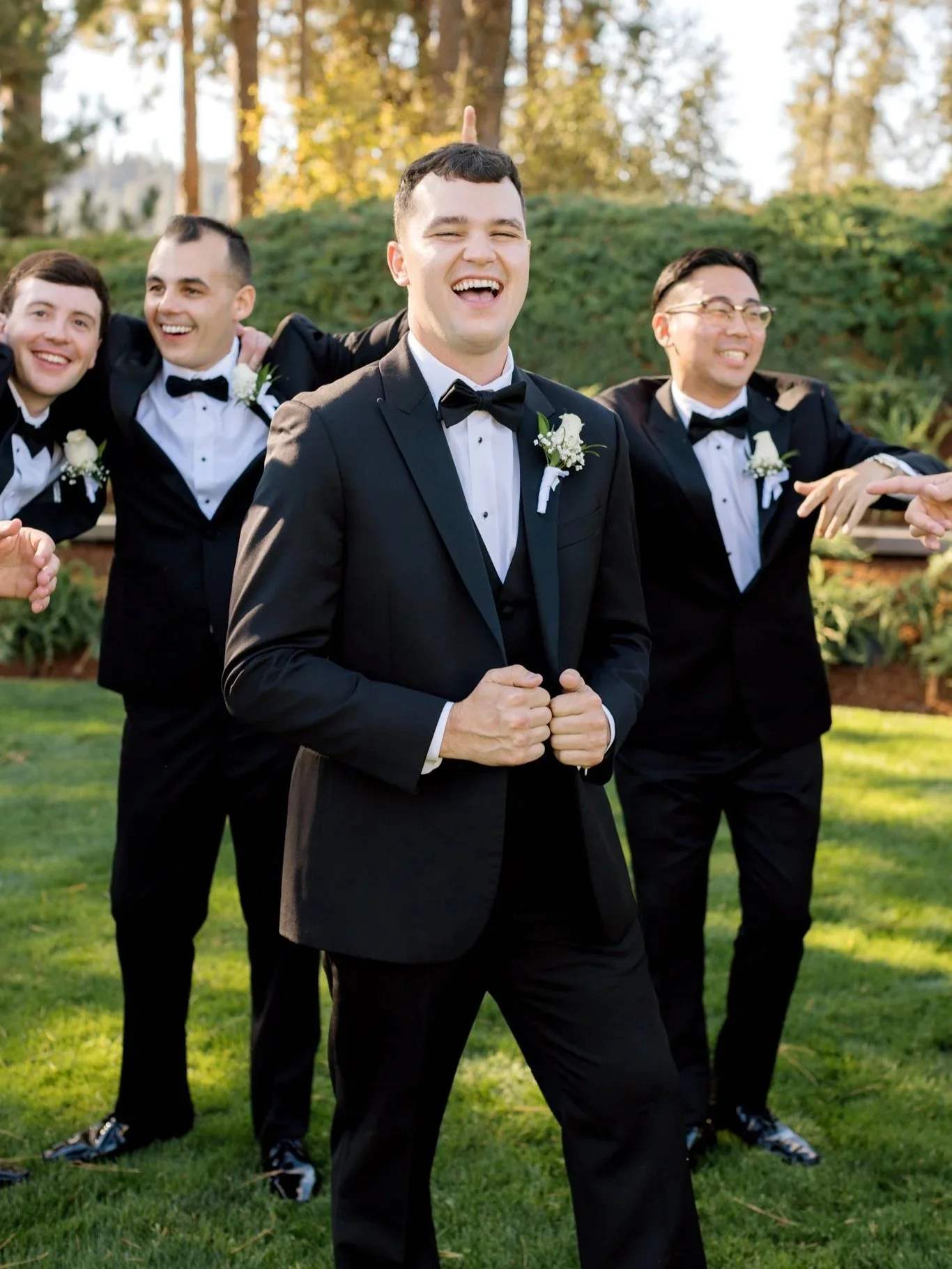 Groom laughing with his groomsmen in black tuxedos during fun wedding party portraits on the lawn in Coeur d’Alene.