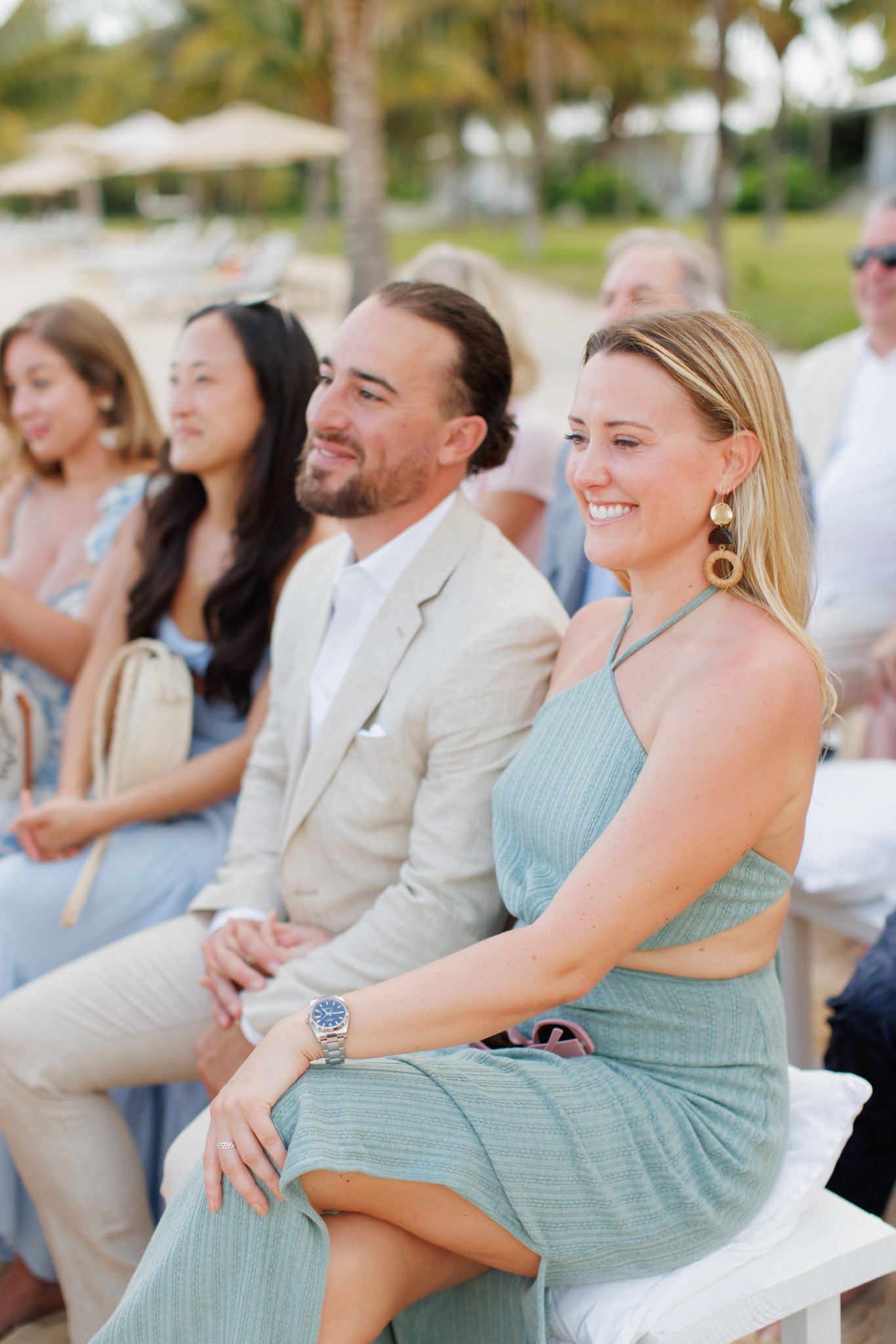 People dressed in light-colored clothing, sitting outdoors, smiling, with some wearing sunglasses, at what appears to be a beachside event or wedding during the daytime.
