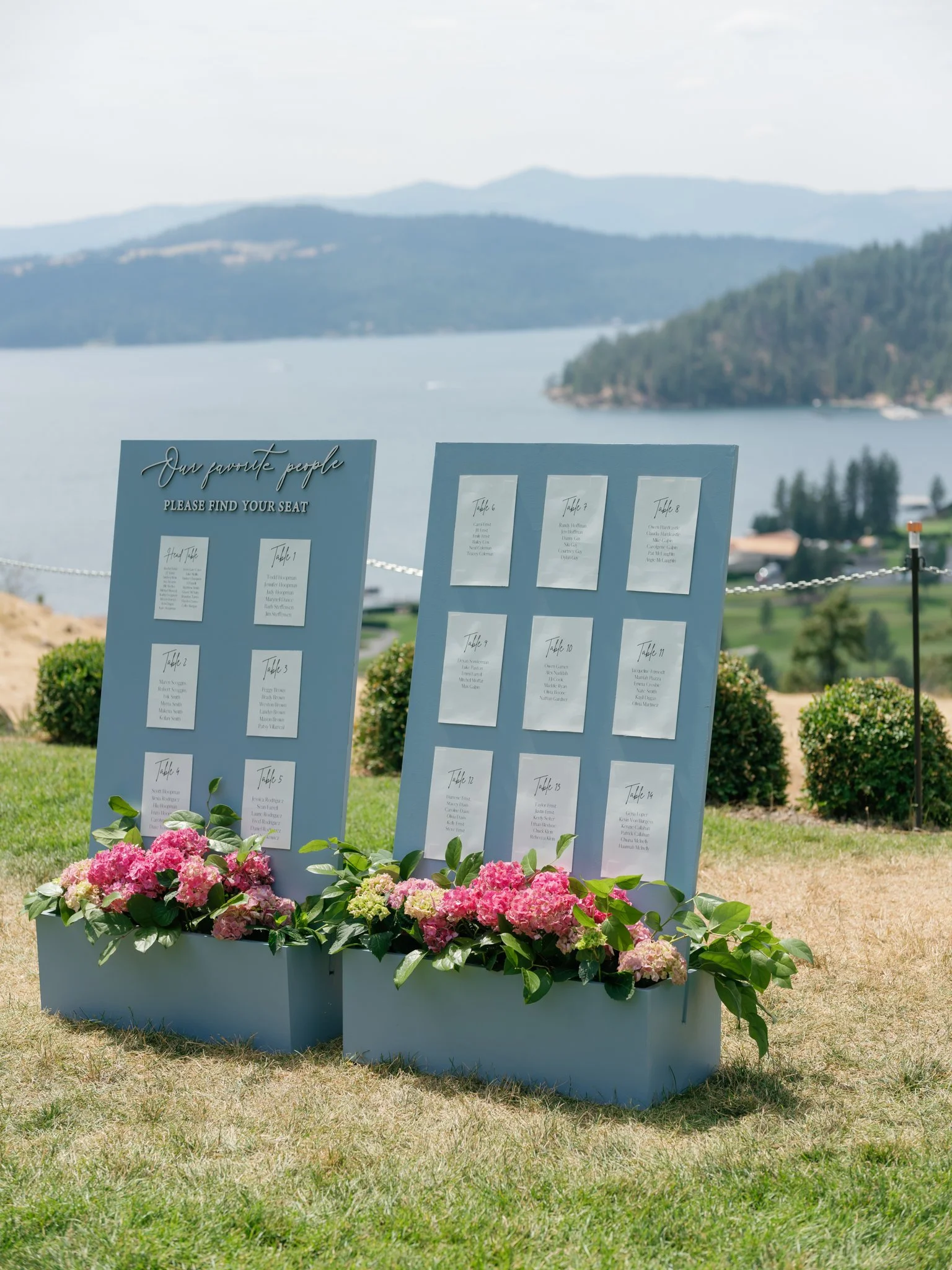 Wedding seating chart with blue boards displaying table assignments, decorated with pink and green floral arrangements, overlooking a lake with mountains in the background.