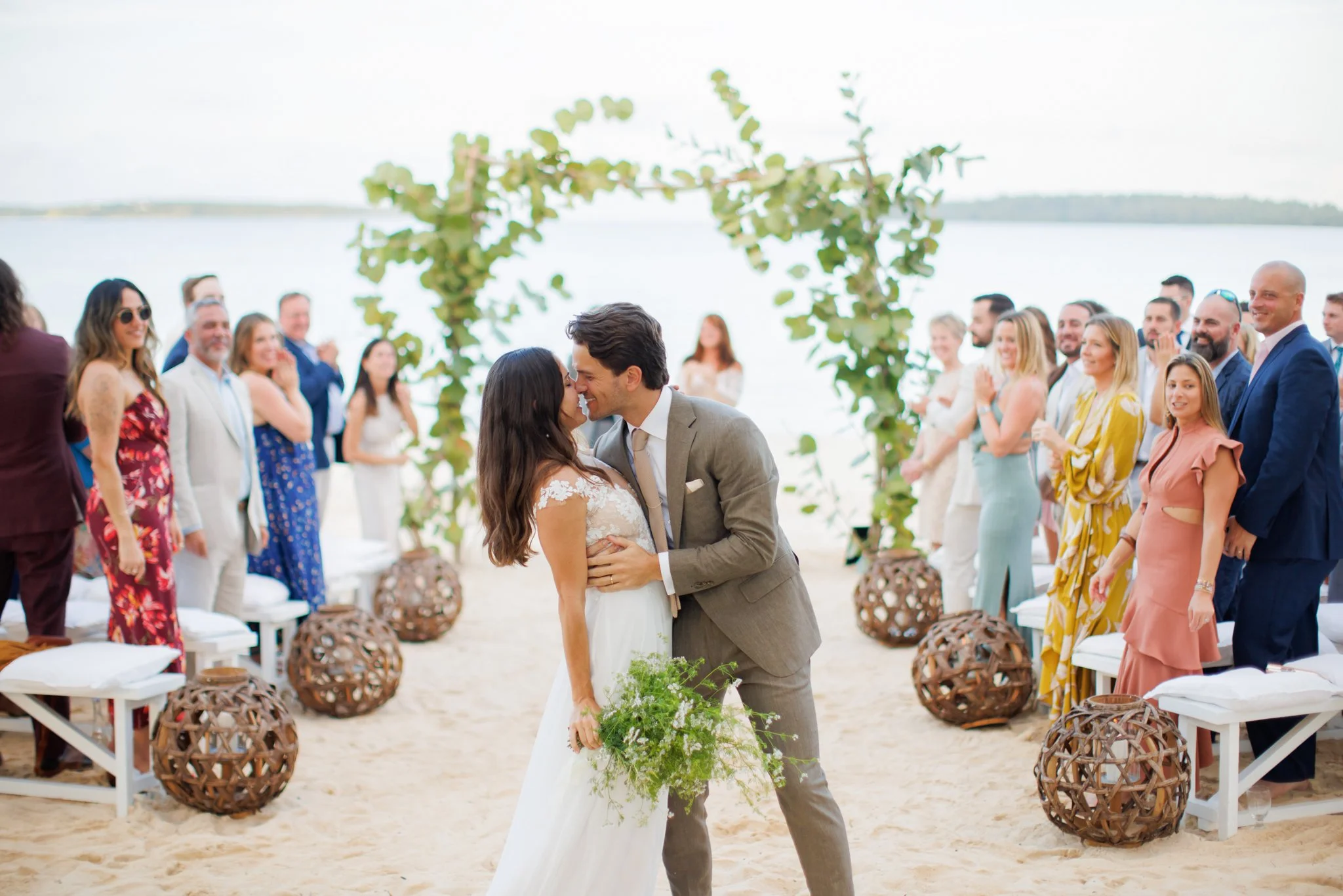A couple is kissing during their beach wedding ceremony, surrounded by friends and family, with a decorative arch and the ocean in the background.