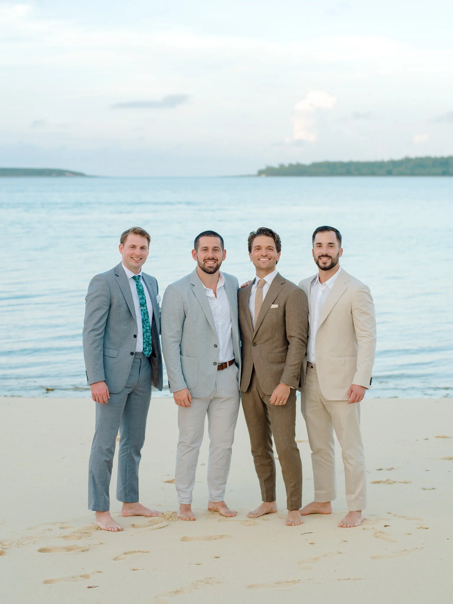 Four men in suits standing barefoot on a beach, smiling with water and sky in the background.