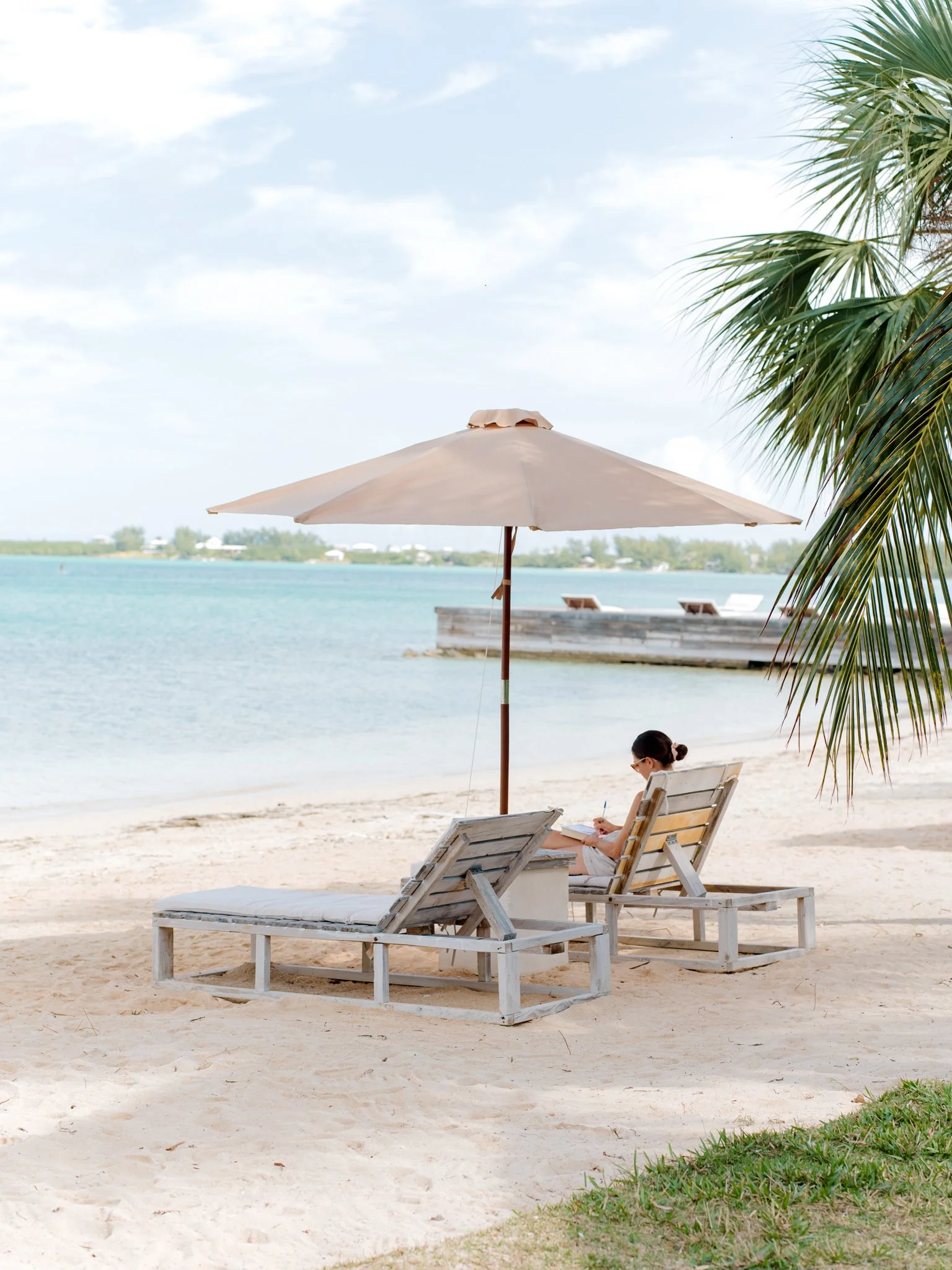 A person sitting on a lounge chair under a large beach umbrella on a sandy beach, reading or writing in a notebook with the ocean and a pier in the background.