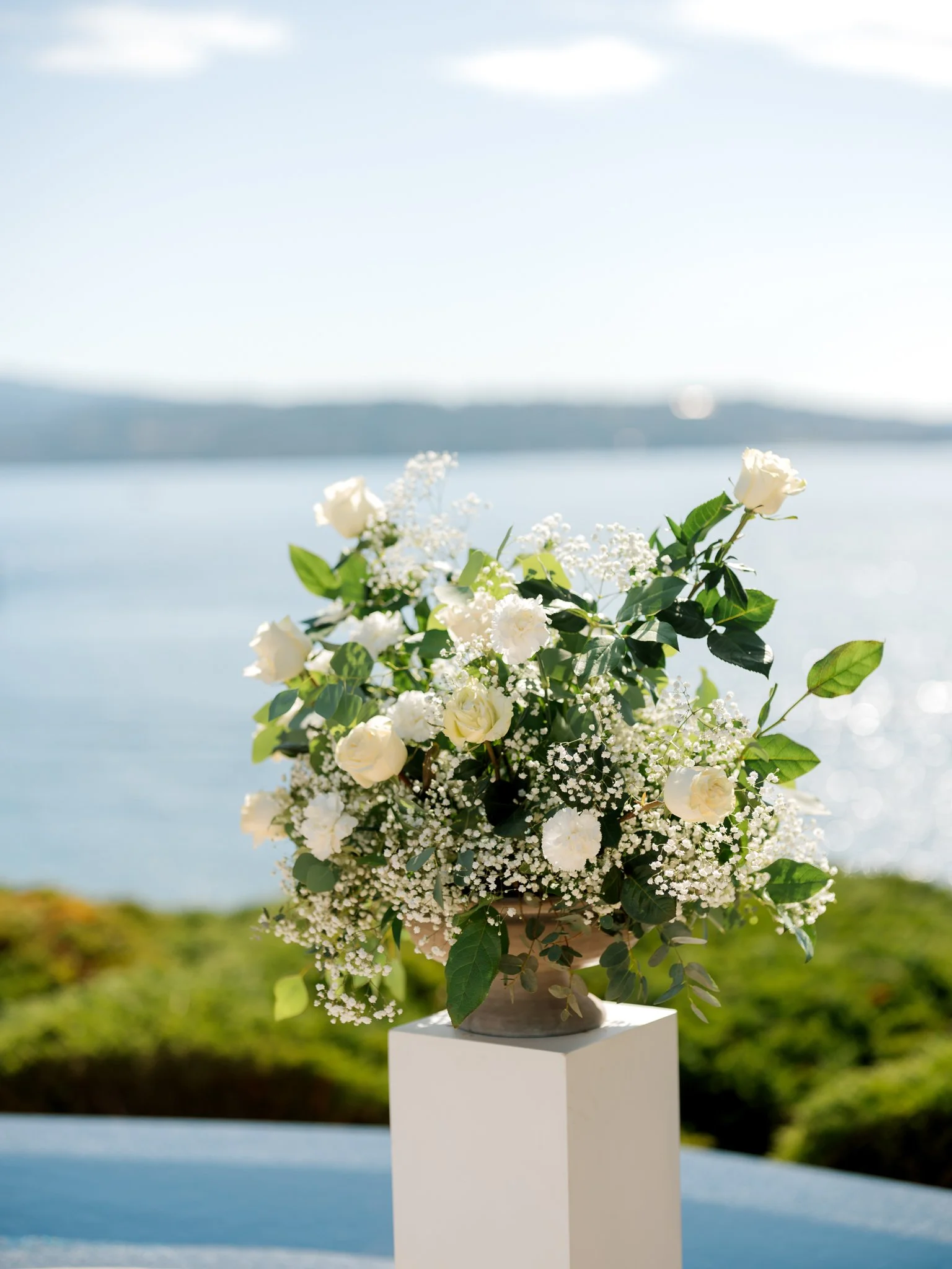 White floral arrangement with roses and greenery displayed on a pedestal overlooking Lake Coeur d’Alene at a lakeside wedding.