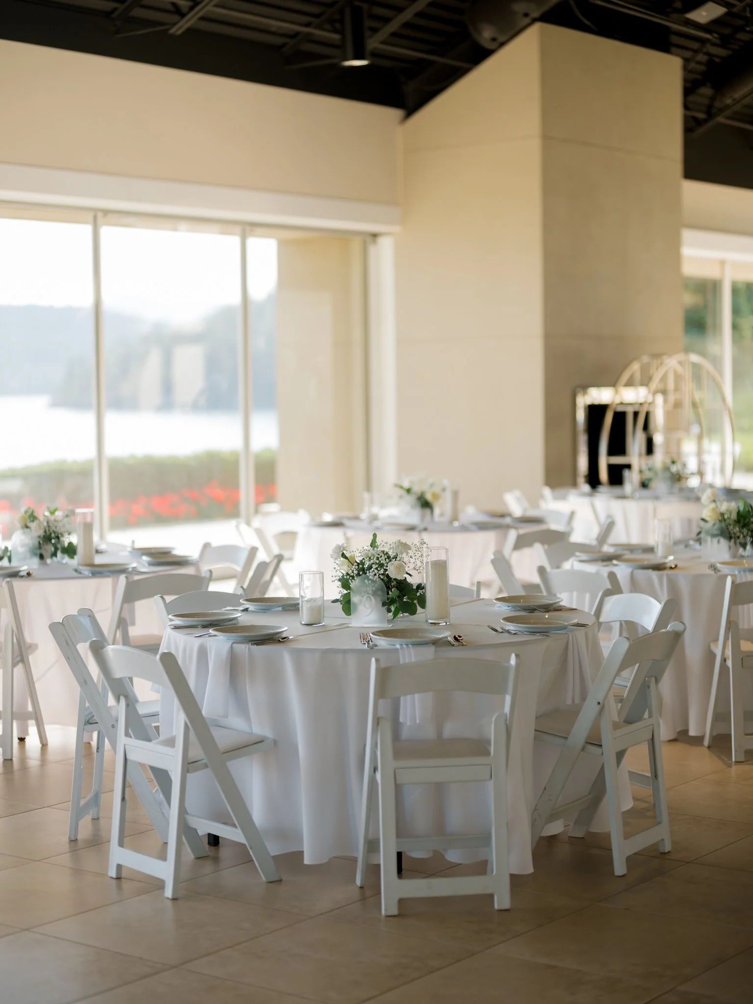 Elegant banquet hall with round tables covered with white tablecloths, set with white plates, glasses, and silverware, and decorated with white floral centerpieces, in front of large windows overlooking a water view.