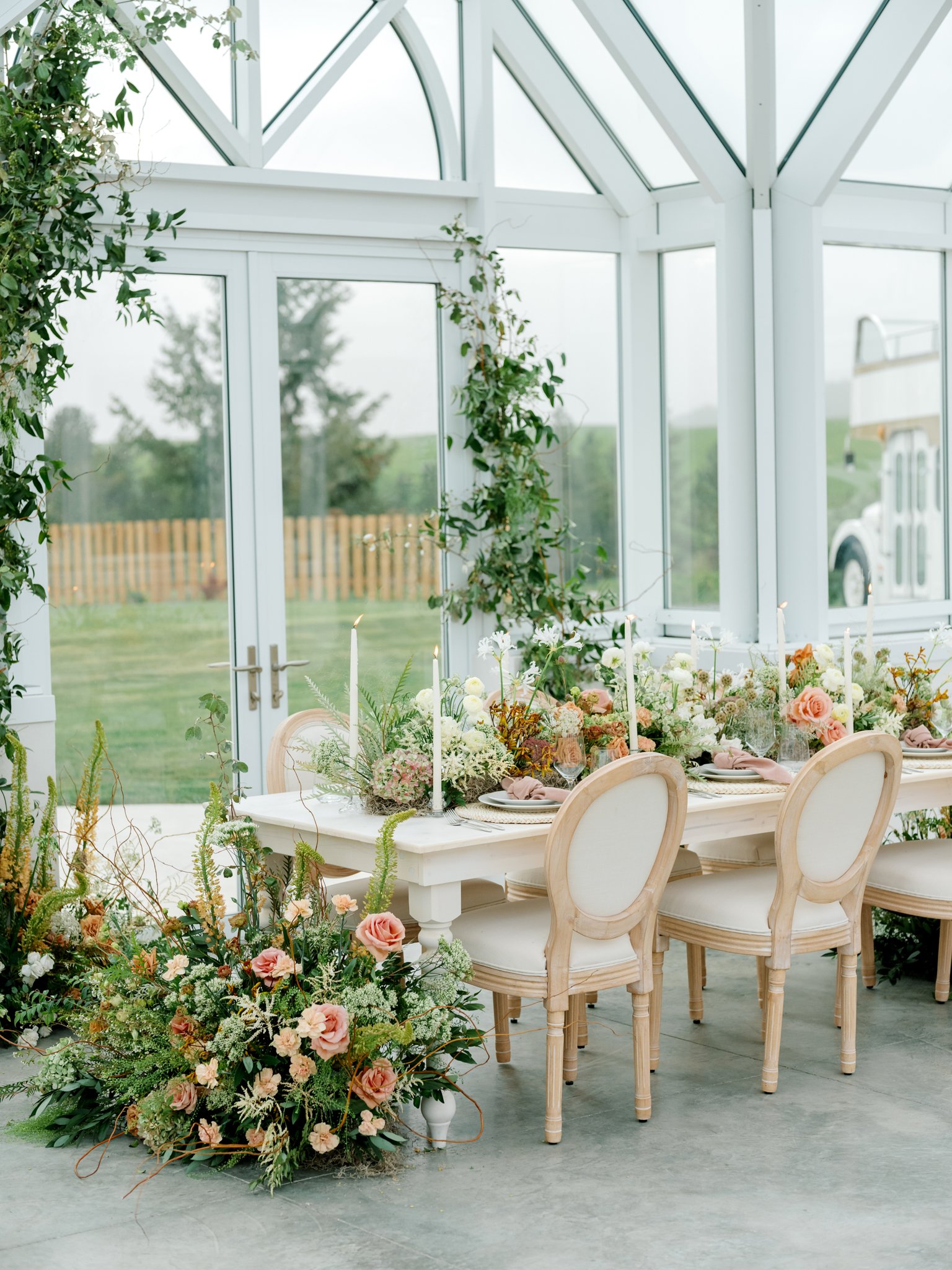 A floral wedding or event table setup inside a bright conservatory with large windows, white furniture, and a lush green outdoor background at Greenhouse on Harvard near Spokane, Washington.