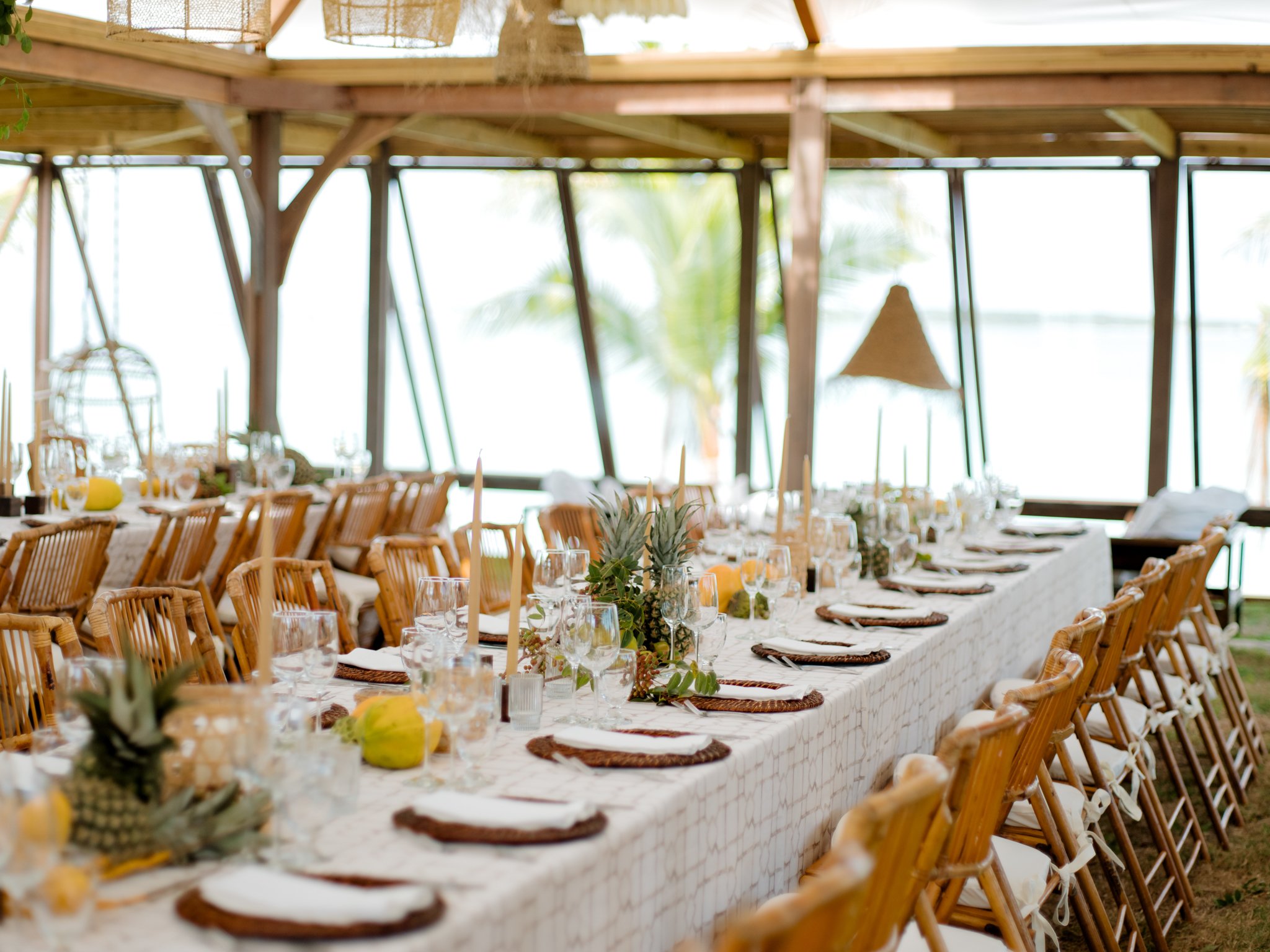 Long dining table decorated with tropical fruits, pineapple and coconuts, set with glassware, plates, napkins, candles, and greenery in a tropical open-air pavilion with wooden beams and large windows.