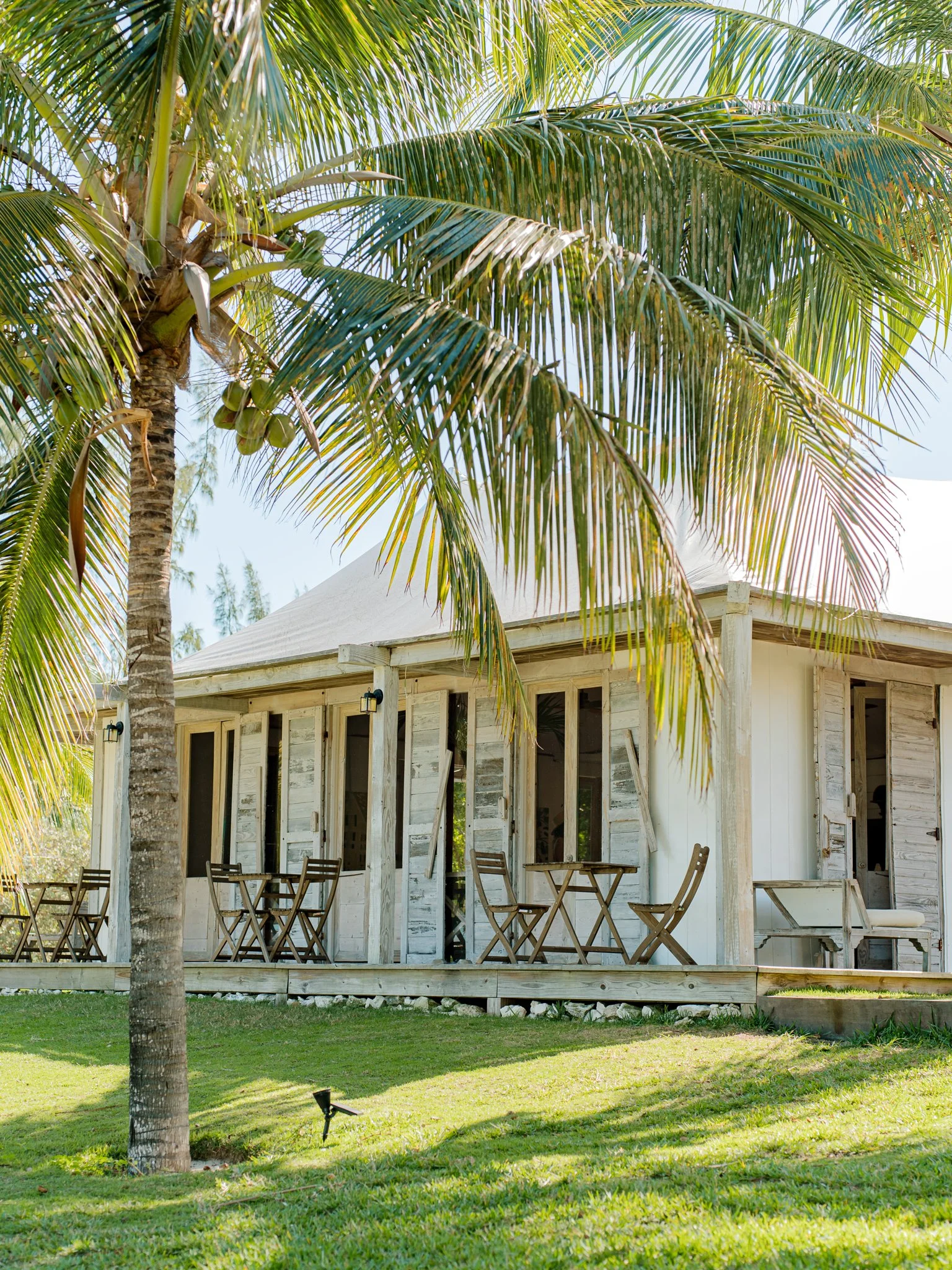 A tropical bungalow with a wooden porch and outdoor tables, surrounded by lush green grass and tall palm trees at The Other Side Hotel during their destination wedding in Eleuthera, Bahamas.
