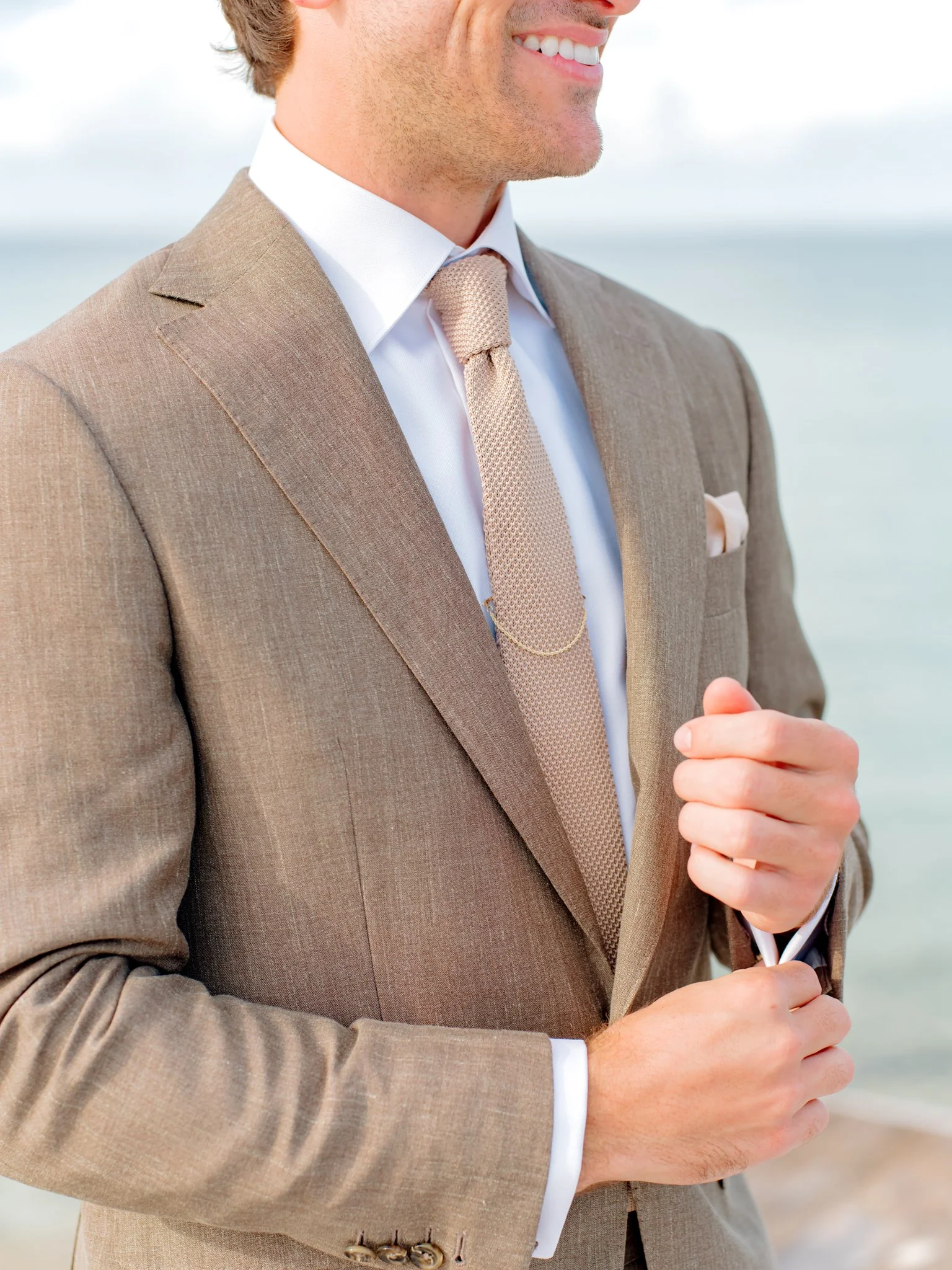 Close-up of a man in a beige suit with a white shirt and beige tie, smiling, standing outdoors near the water, adjusting his suit cuff.