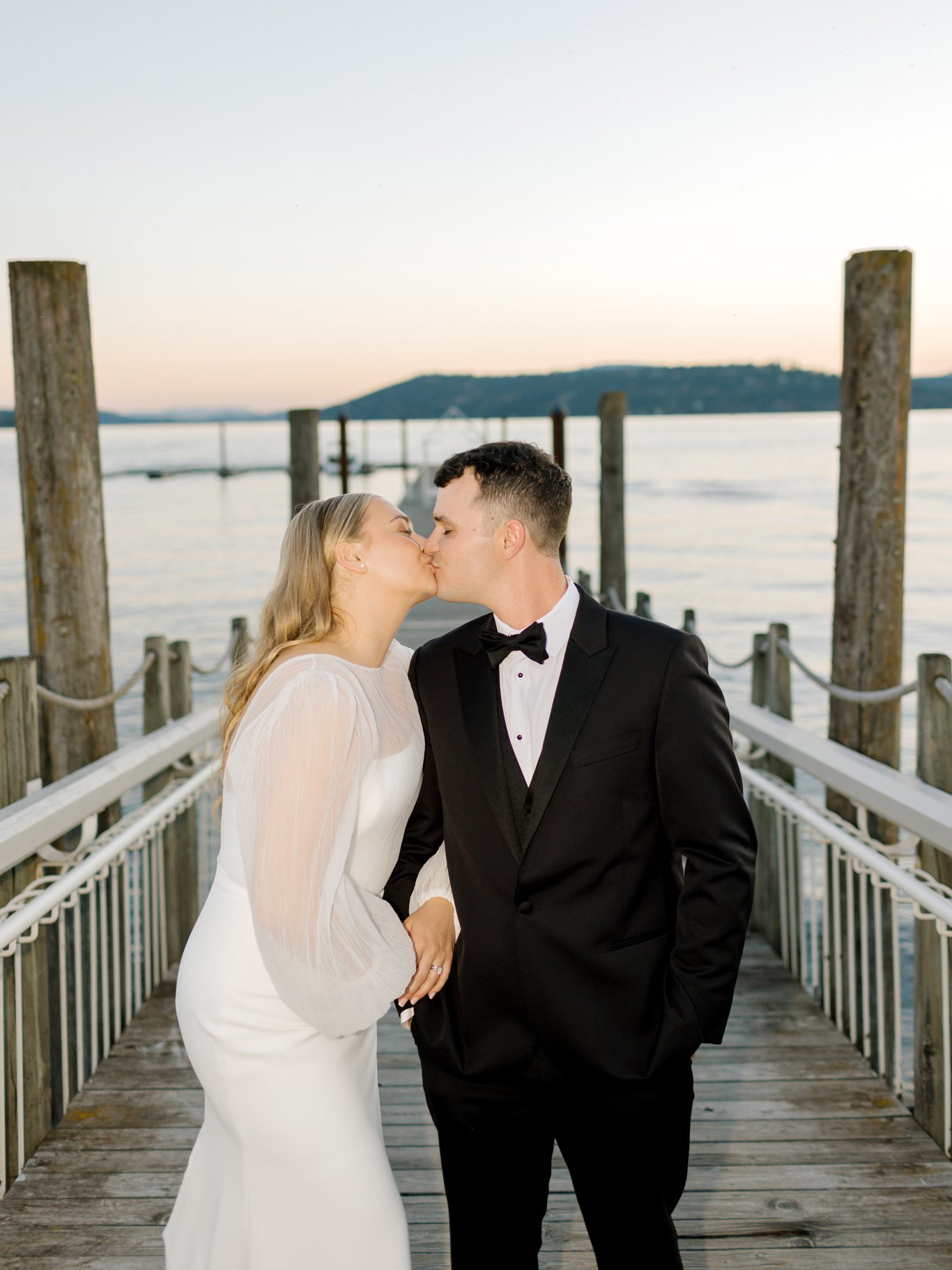 A newlywed couple wearing wedding attire kiss on a wooden dock with water and hills in the background at sunset at the Hagadone Event Center in Coeur d'Alene, Idaho.