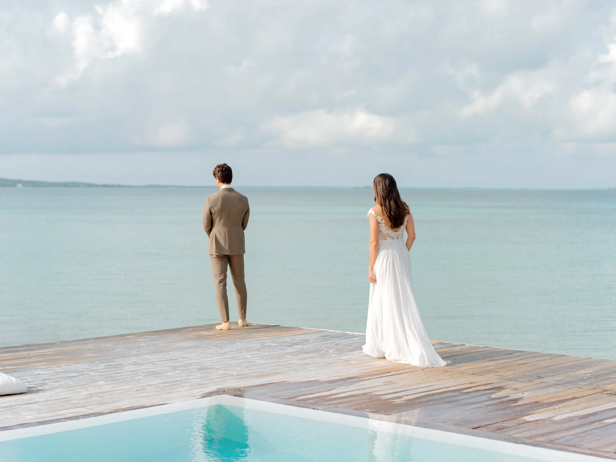 A man and woman in wedding attire standing on a wooden dock, facing the ocean on a partly cloudy day.