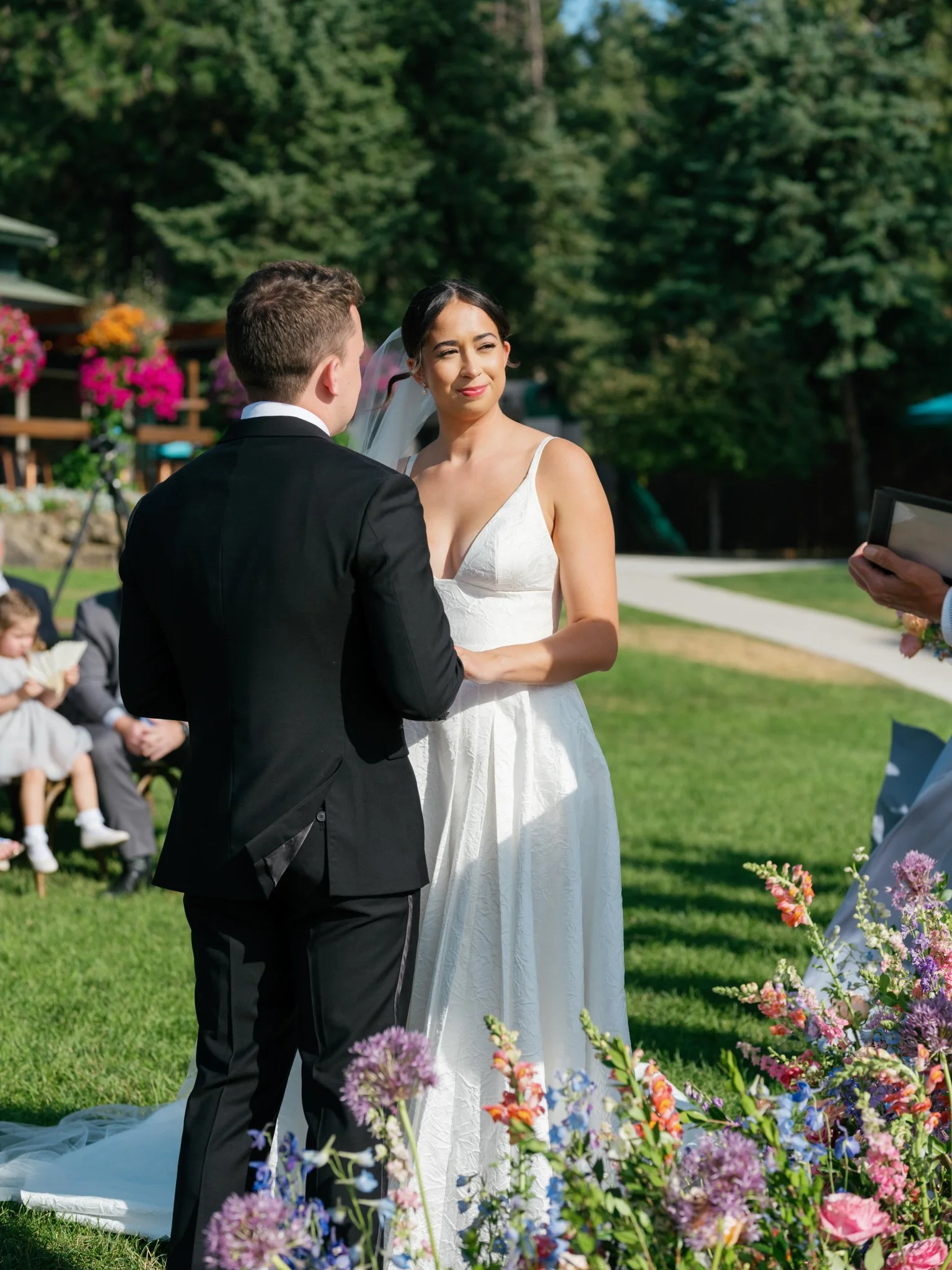 A bride and groom holding hands during an outdoor wedding ceremony, with guests sitting on the grass in the background and colorful flowers in the foreground.