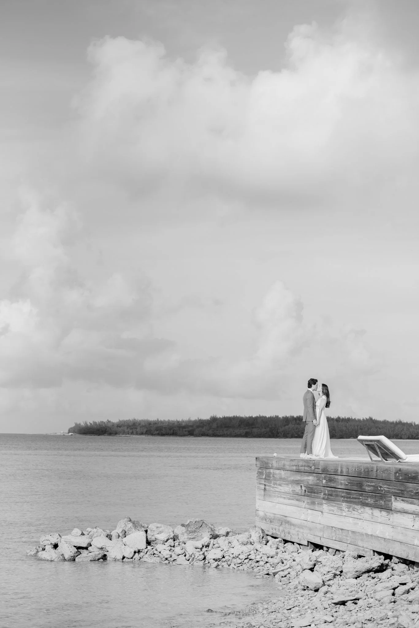 A bride and groom standing on a wooden dock by the water, sharing a kiss, under cloudy skies at The Other Side Hotel Eleuthera during a luxury Bahamas destination wedding.