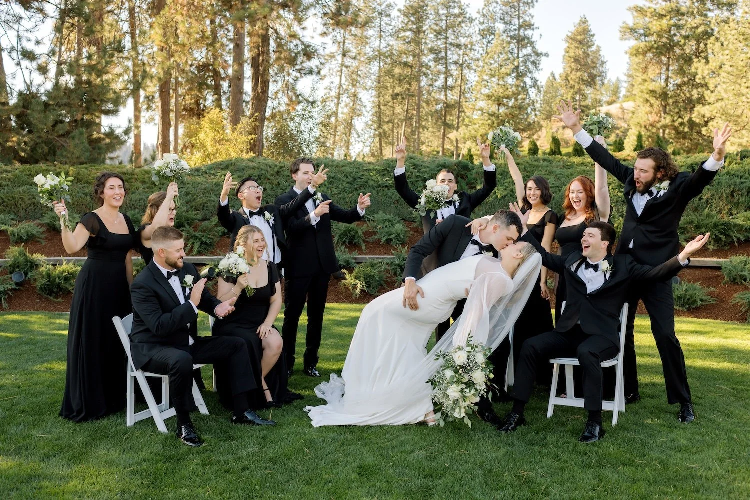 A group of wedding party members dressed in formal black attire celebrating outdoors, with the bride and groom kissing in the center, surrounded by friends who are holding bouquets, raising hands, and smiling.