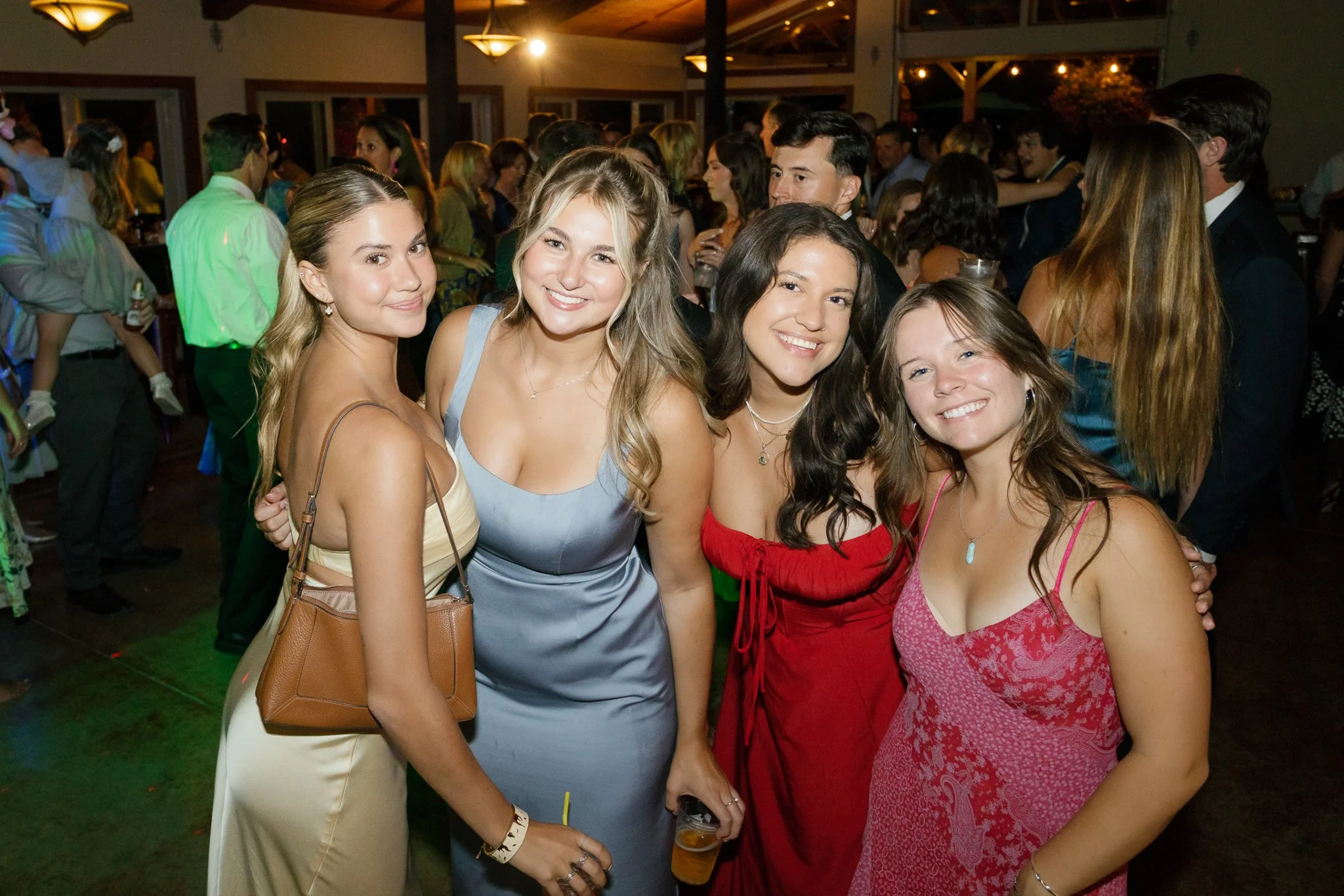 Four women smiling at a social gathering or party, standing close together, with other people in the background.