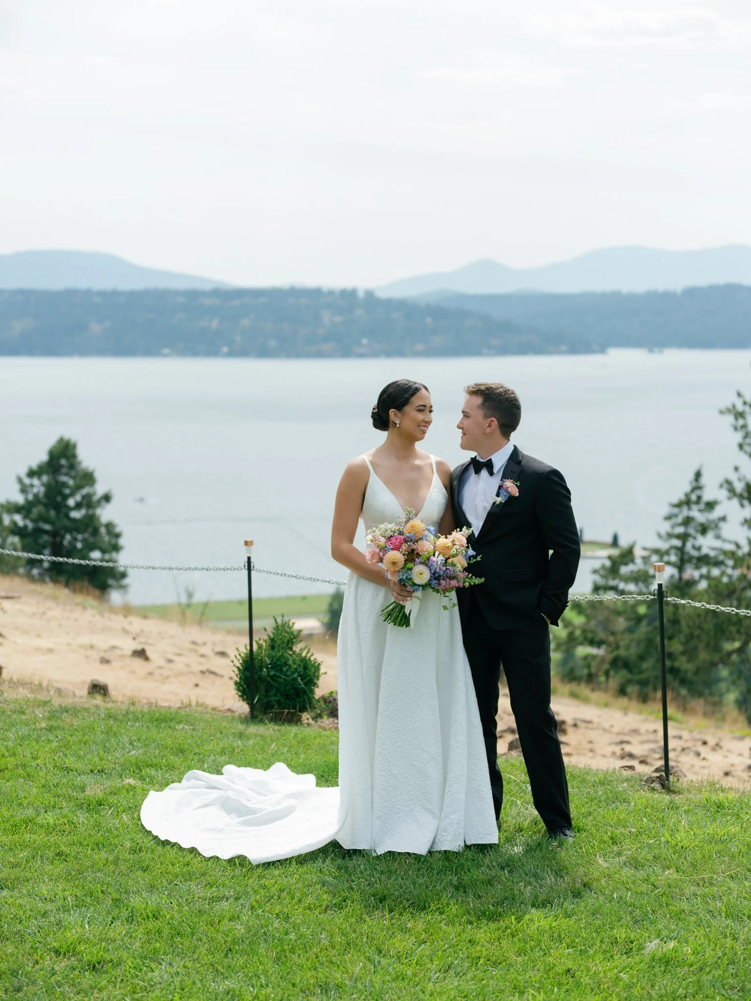 Bride and groom standing on green grass near water with mountains in background, smiling at each other, bride holding a colorful bouquet, outdoor wedding scene at Elk Point wedding above Lake Coeur d’Alene.