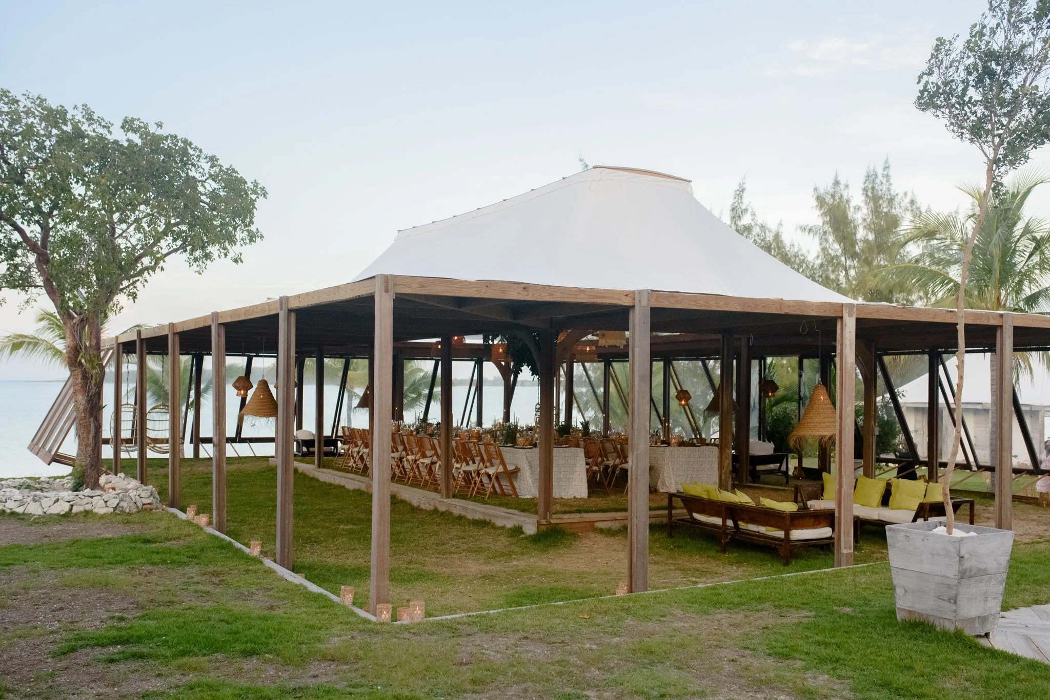 An outdoor event space with a large open-sided wooden structure topped with a white canopy, surrounded by trees and greenery, with a view of the water in the background, decorated with hanging lamps and yellow cushions.