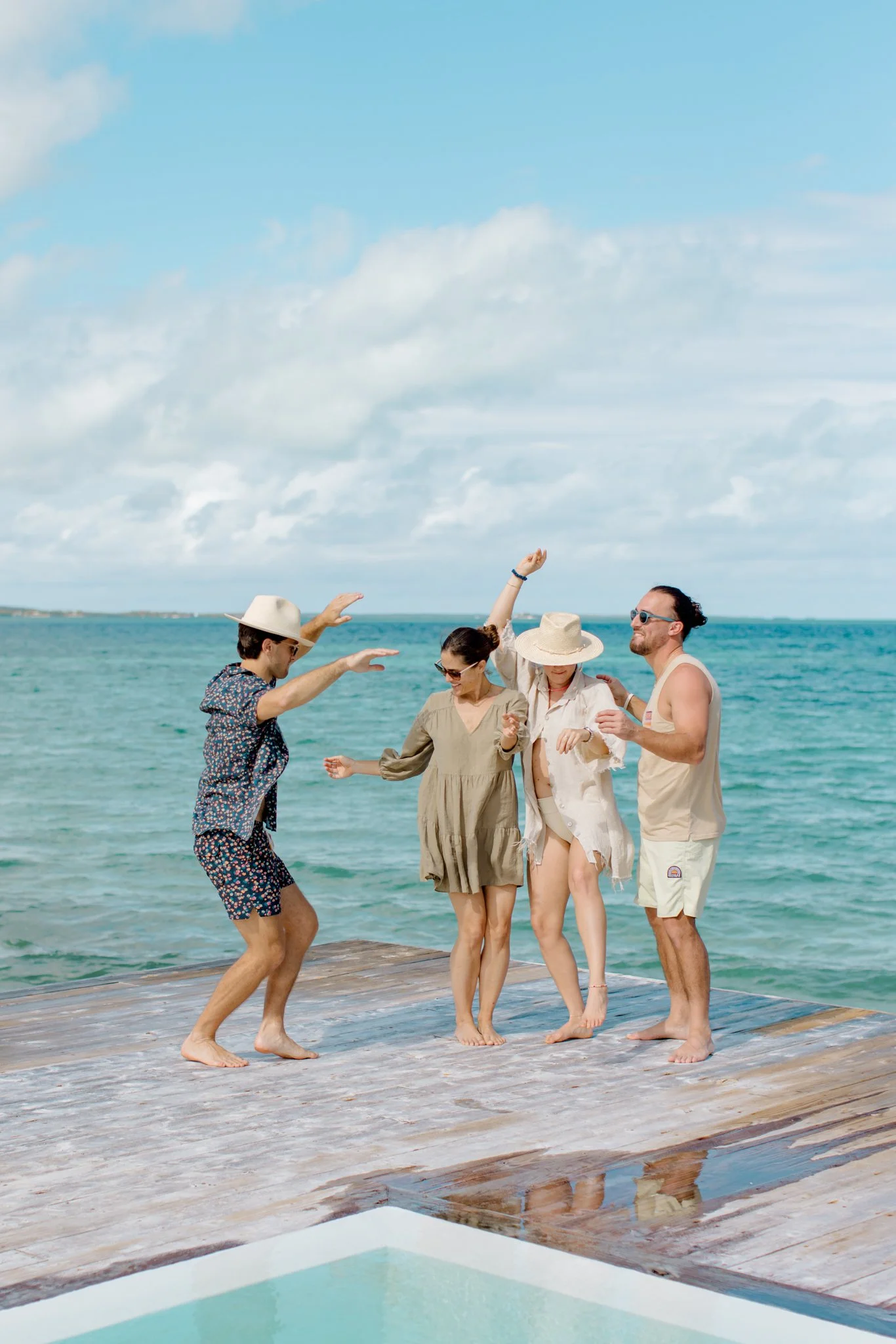 Four friends dancing and having fun on a wooden dock by the ocean on a sunny day, dressed in casual summer clothes and hats at The Other Side Hotel during their destination wedding in Eleuthera, Bahamas.