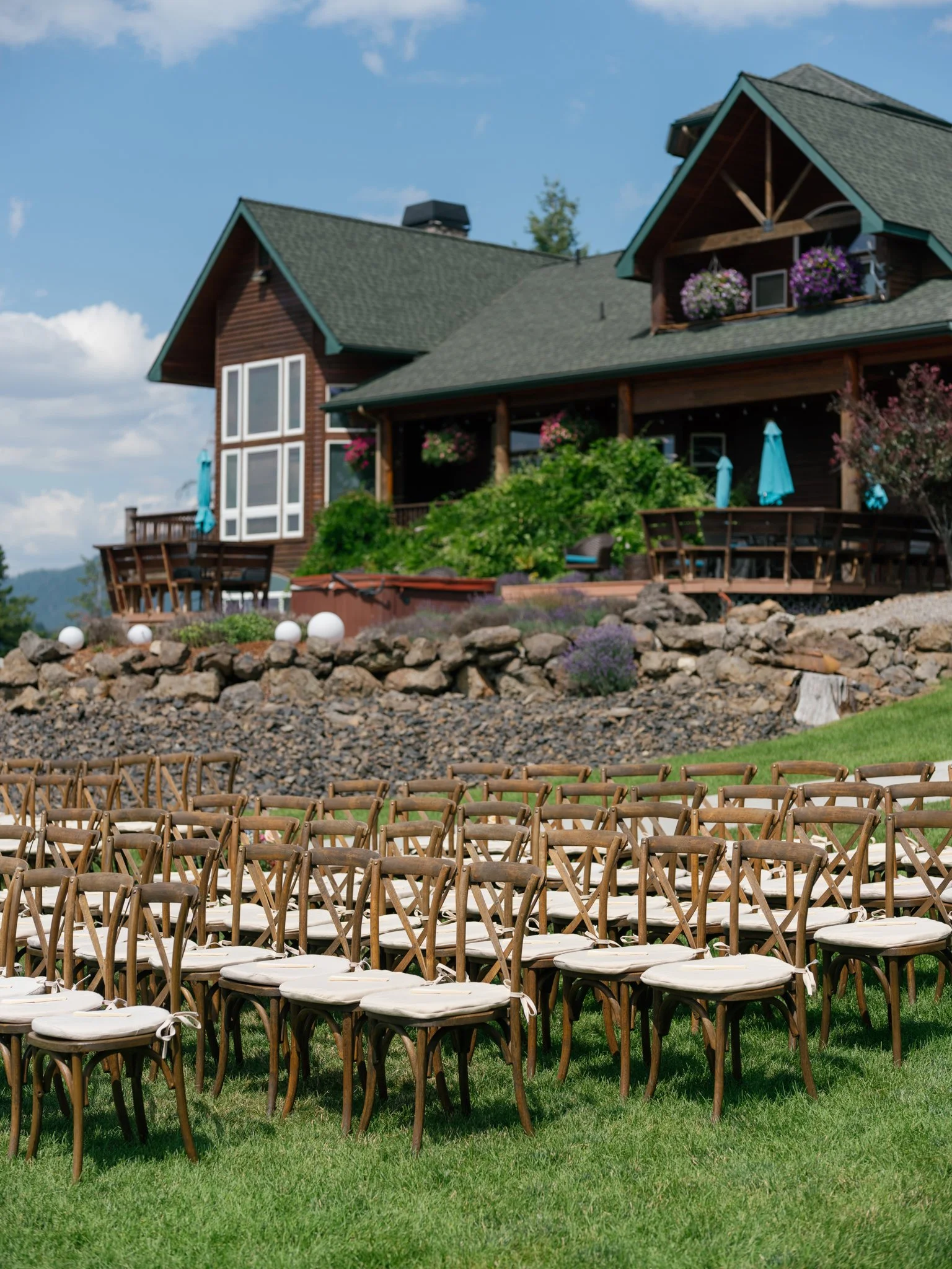 An outdoor wedding setup with rows of chairs on a grassy lawn, facing a large wooded house with a deck and colorful umbrellas, under a partly cloudy sky at Elk Point wedding above Lake Coeur d’Alene.