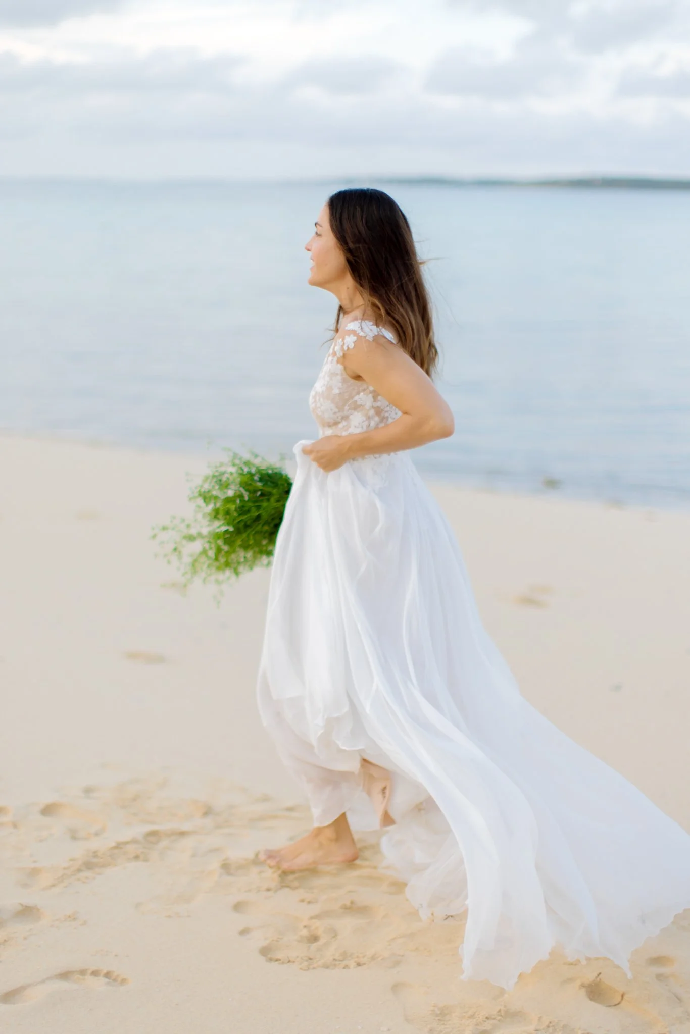 Woman in a white dress walking barefoot on a sandy beach near the water, holding a bouquet of green flowers.