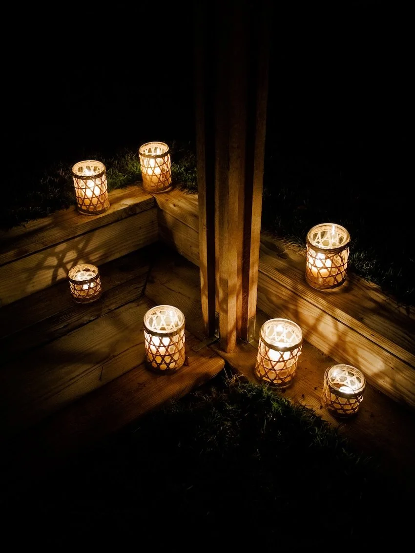Seven lit candles in woven holders placed on a wooden structure outdoors at night.