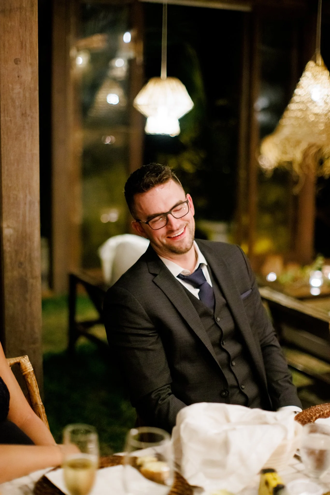 A man in a suit and glasses smiling at a dinner party in a warmly lit room.