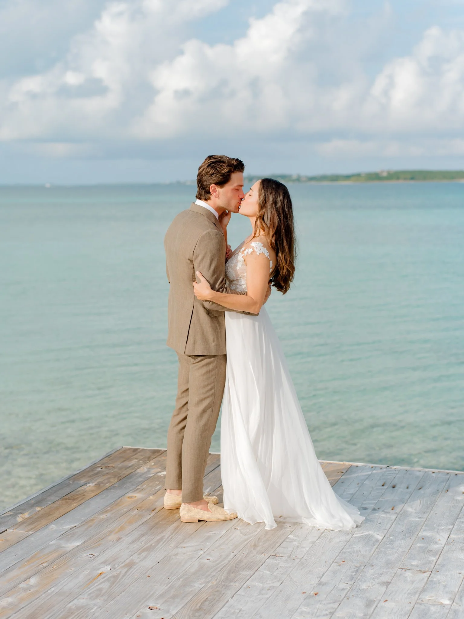 A couple dressed in wedding attire standing on a wooden dock by the water, about to kiss, with cloudy sky and distant shoreline in the background at The Other Side Hotel Eleuthera during a luxury Bahamas destination wedding.