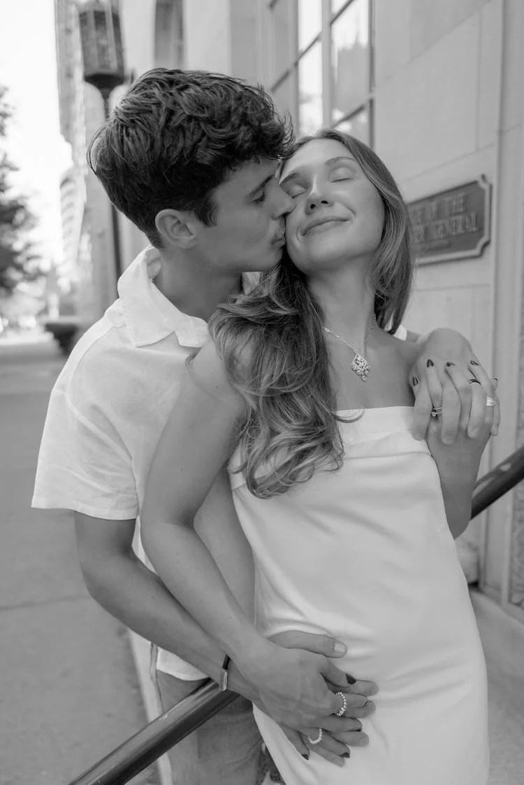 A black and white photo of a couple standing outdoors, with the man kissing the woman's cheek, both wearing light-colored clothing. The woman has long wavy hair and is smiling with her eyes closed, wearing a necklace and rings, and the man has short 