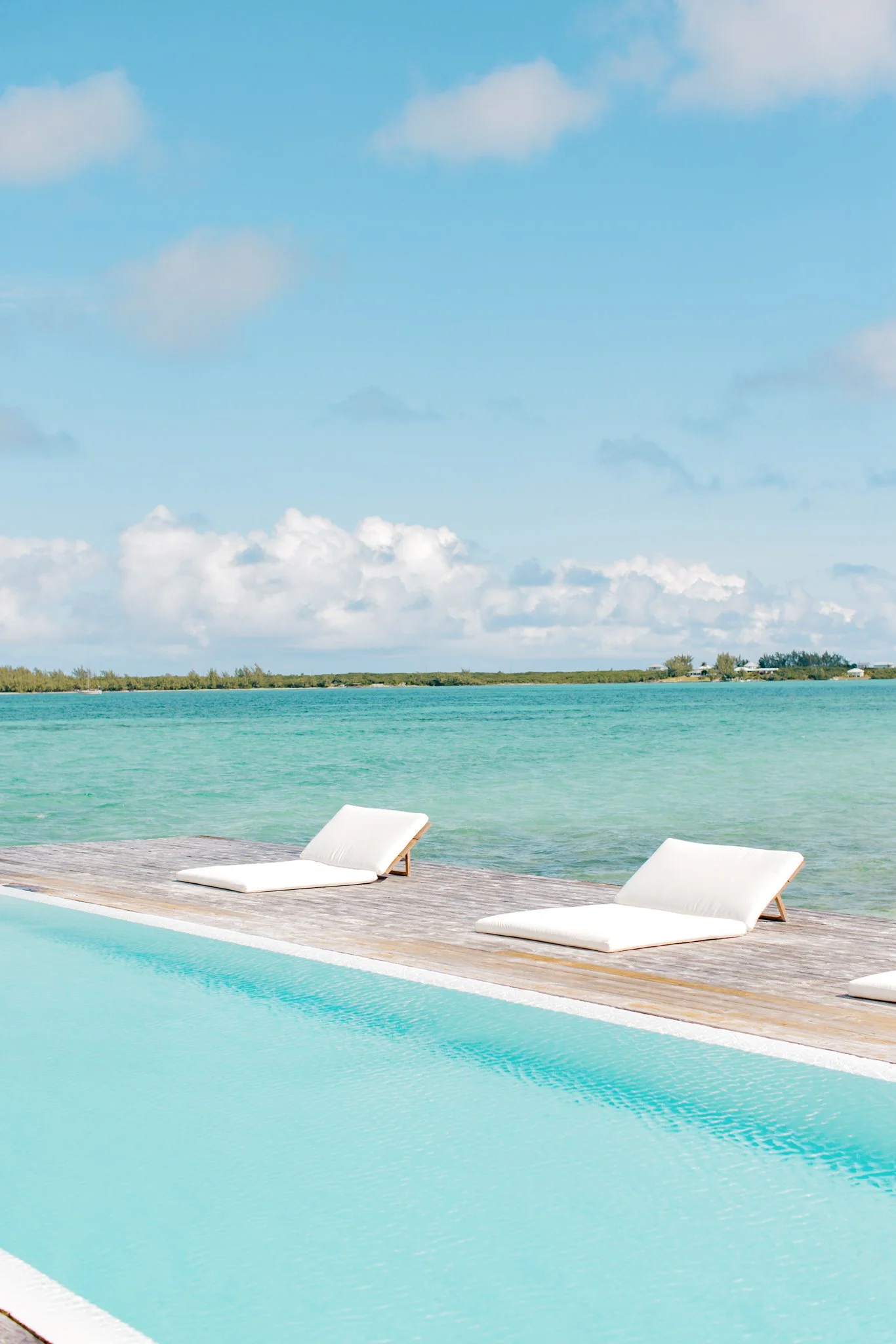 Empty swimming pool with white lounge chairs on a wooden deck overlooking turquoise water and a cloudy blue sky.