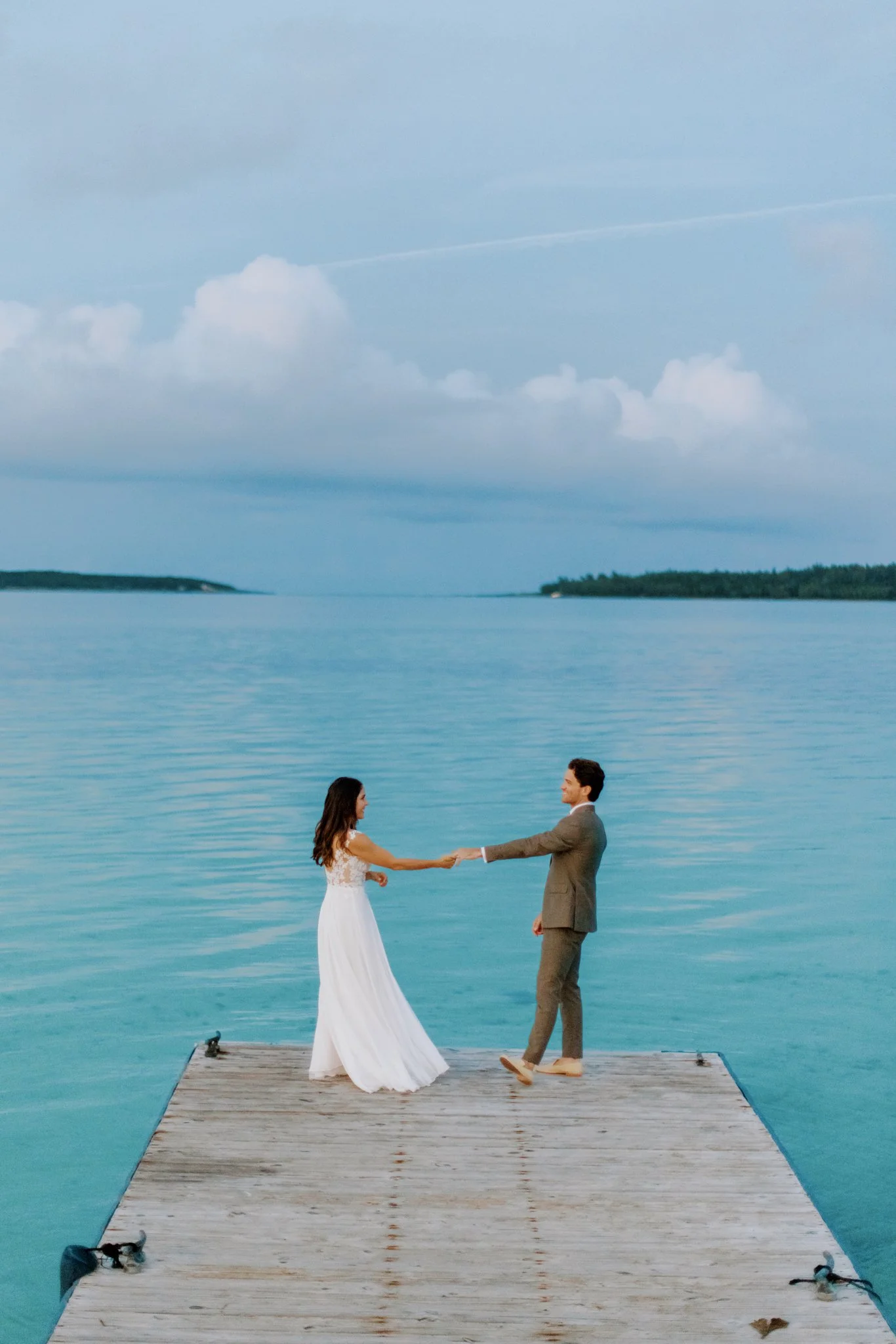 A couple, dressed in wedding attire, holding hands on a wooden dock by a calm, blue lake with a cloudy sky overhead.