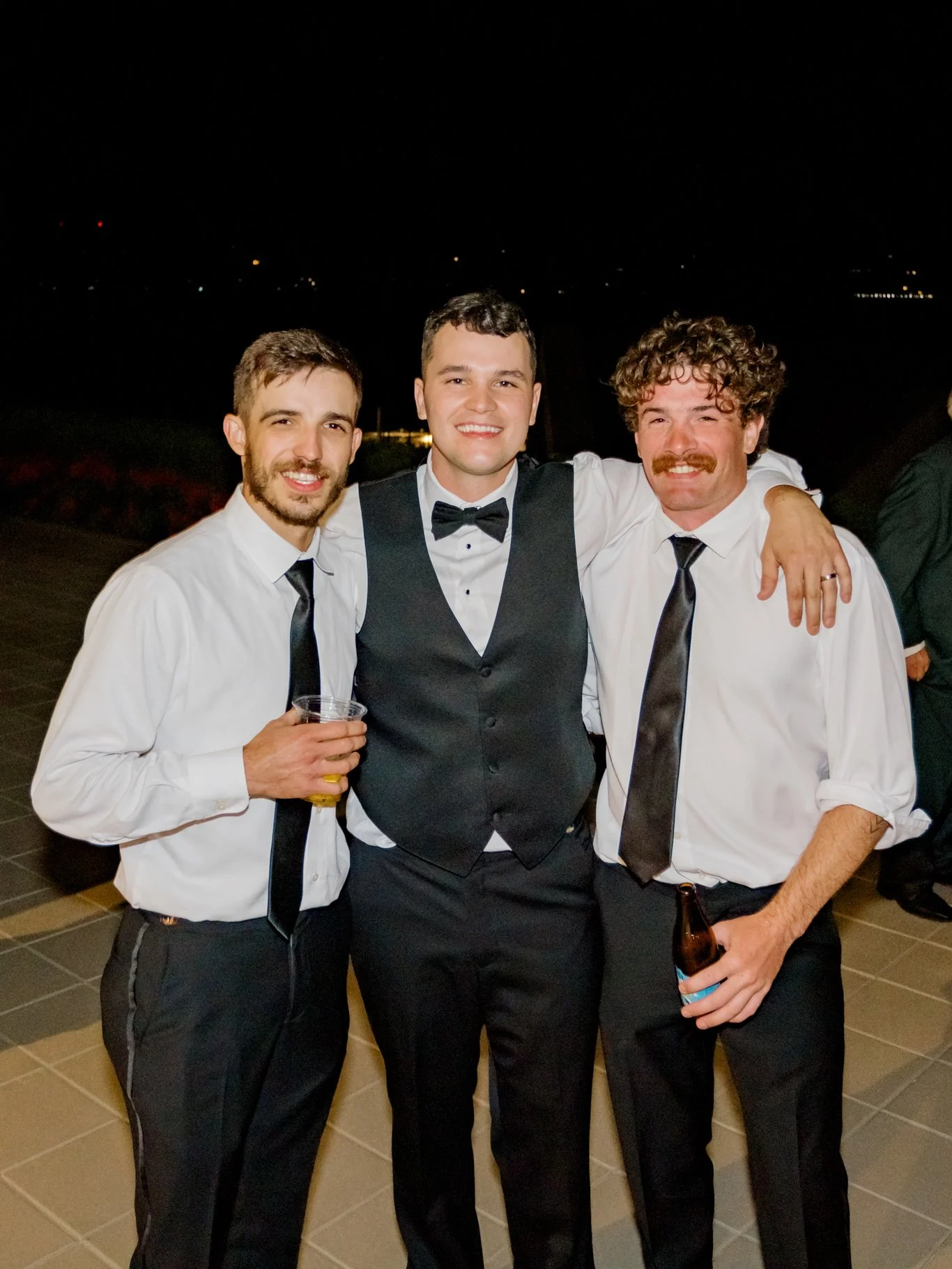 Three men dressed in formal attire at an outdoor evening event, standing close together and smiling at the camera. One man on the left holds a drink, the man in the middle is wearing a tuxedo with a bow tie, and the man on the right holds a beer.