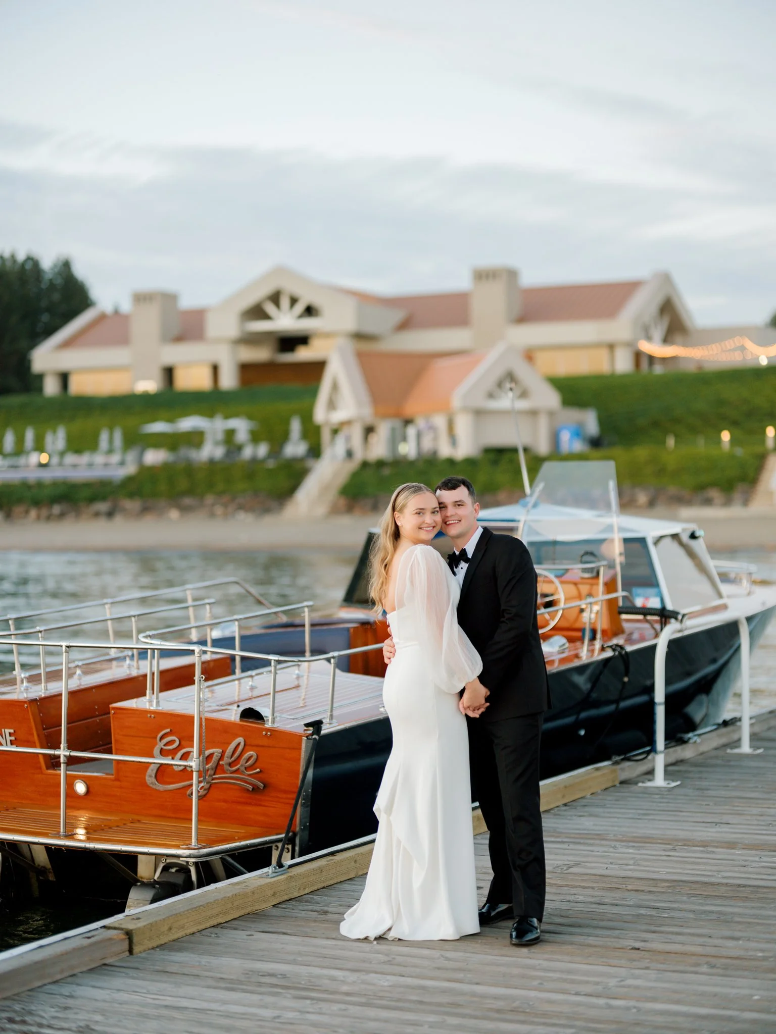 A bride and groom in wedding attire standing on a dock next to a boat with a house in the background.