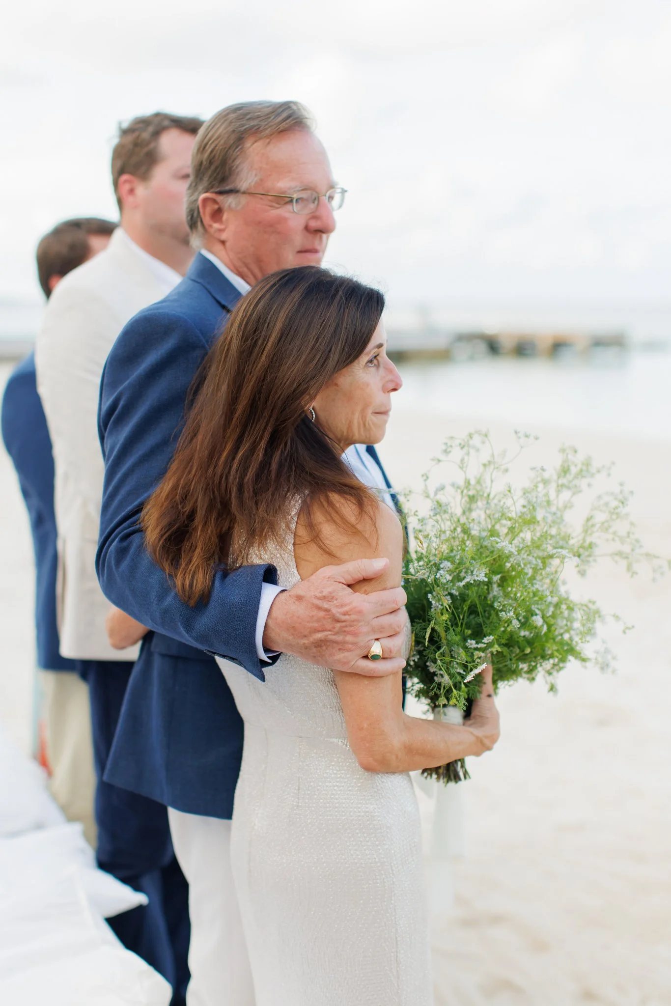 An older man in a blue jacket gently holds a woman in a wedding dress holding a bouquet of greenery during a beach wedding ceremony.