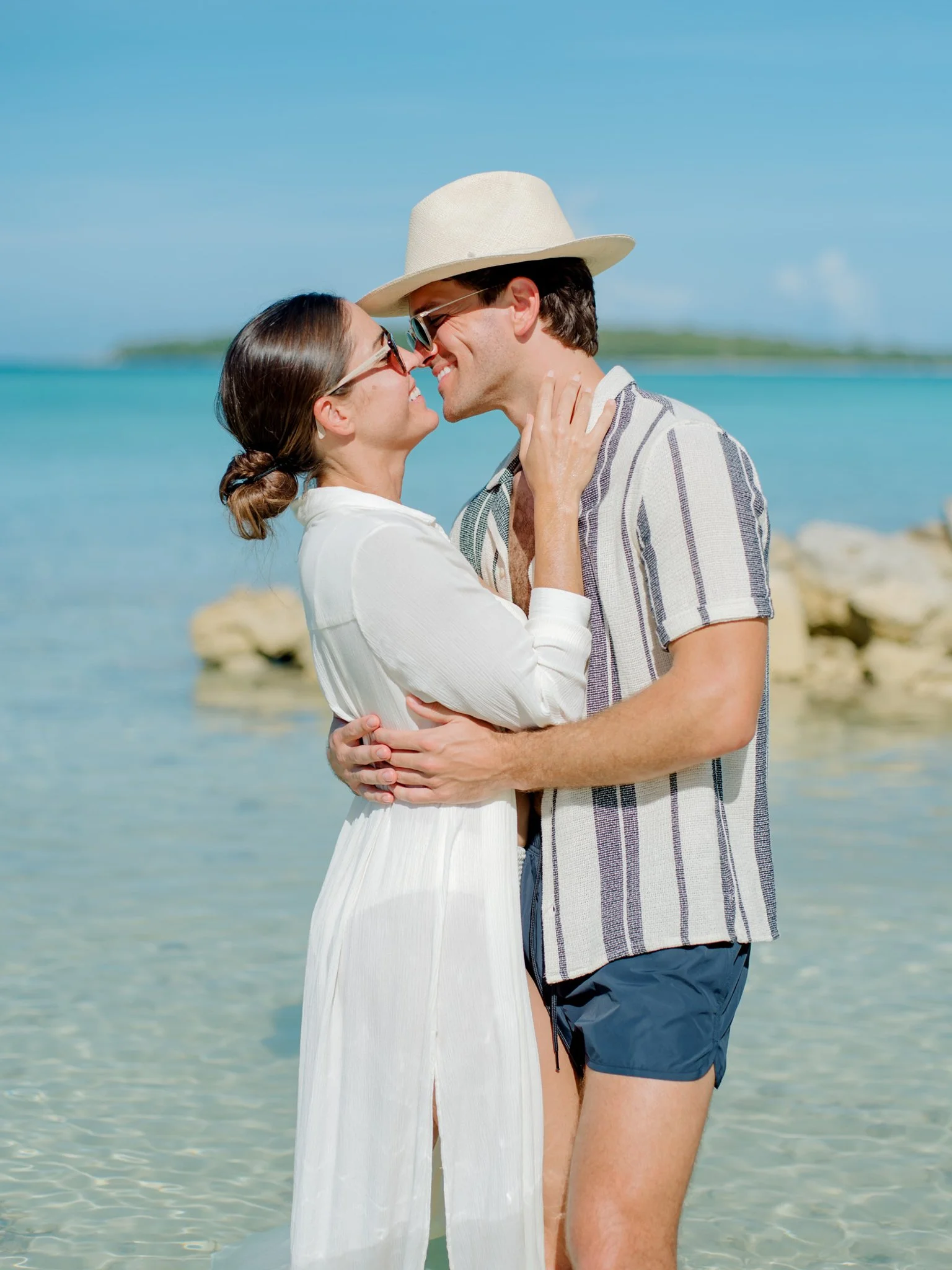 A couple embracing on a beach, smiling and looking into each other's eyes, with the ocean and a small island in the background at The Other Side Hotel Eleuthera during a luxury Bahamas destination wedding.