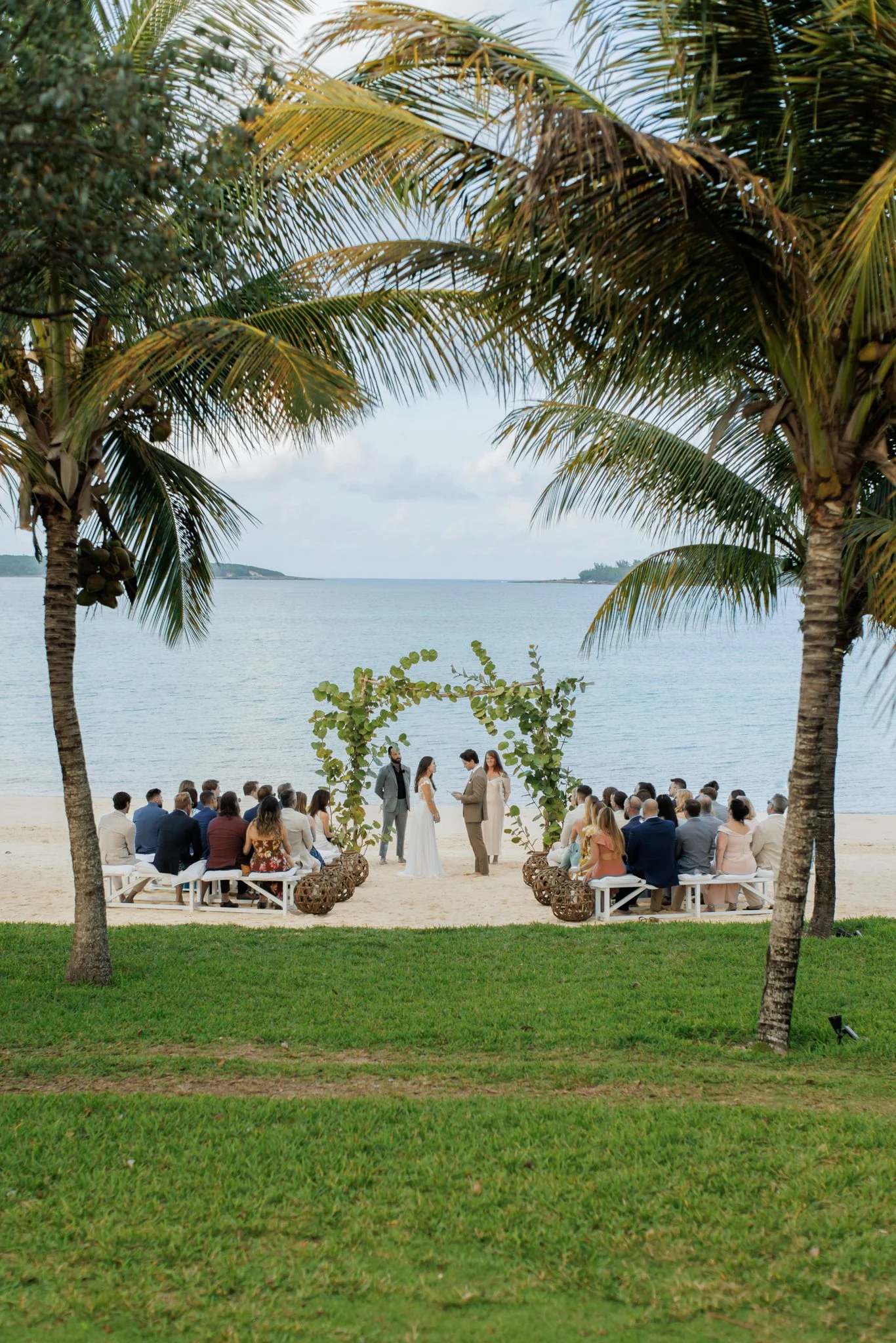 A beach wedding ceremony on a sandy beach with an ocean backdrop, framed by palm trees. Guests are seated on benches, and the couple is exchanging vows under a decorated arch.