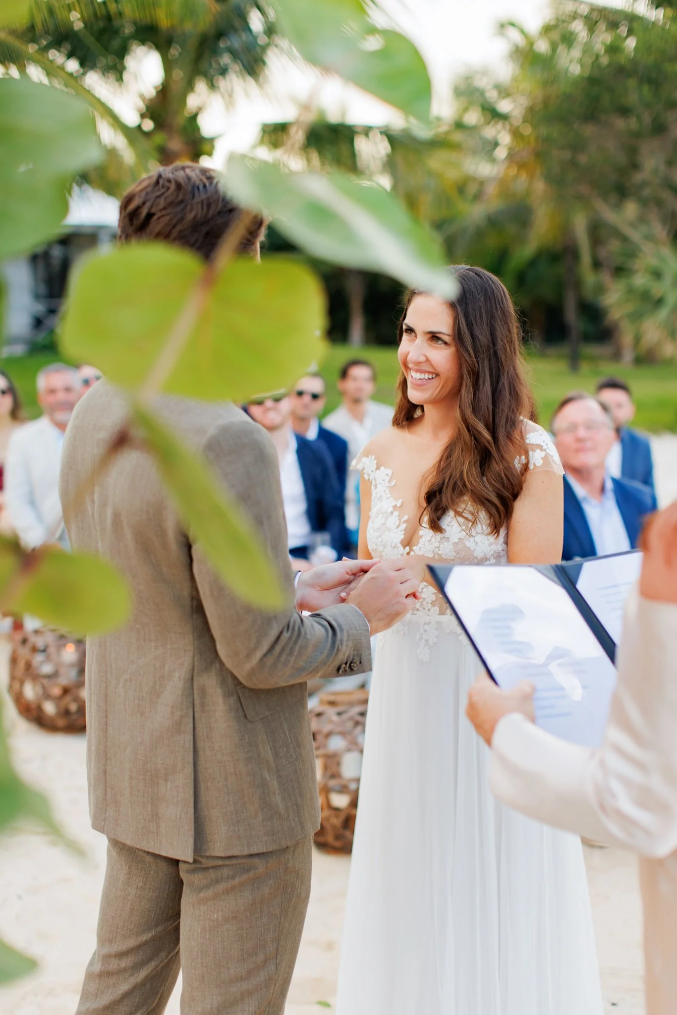A couple stands outdoors at their wedding ceremony, holding hands and smiling at each other, with guests seated behind them, surrounded by greenery and trees.