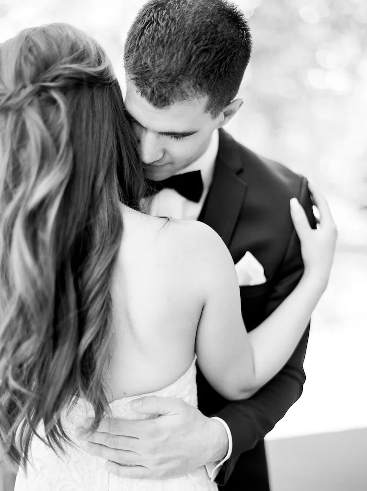 A black and white photo of a couple close together, embracing. The woman has long wavy hair and is wearing a strapless dress. The man is wearing a tuxedo with a bow tie and is gently holding the woman with one hand on her back.